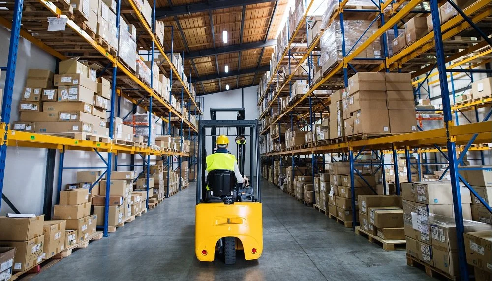 Warehouse worker operating a forklift among shelves stacked with boxes.