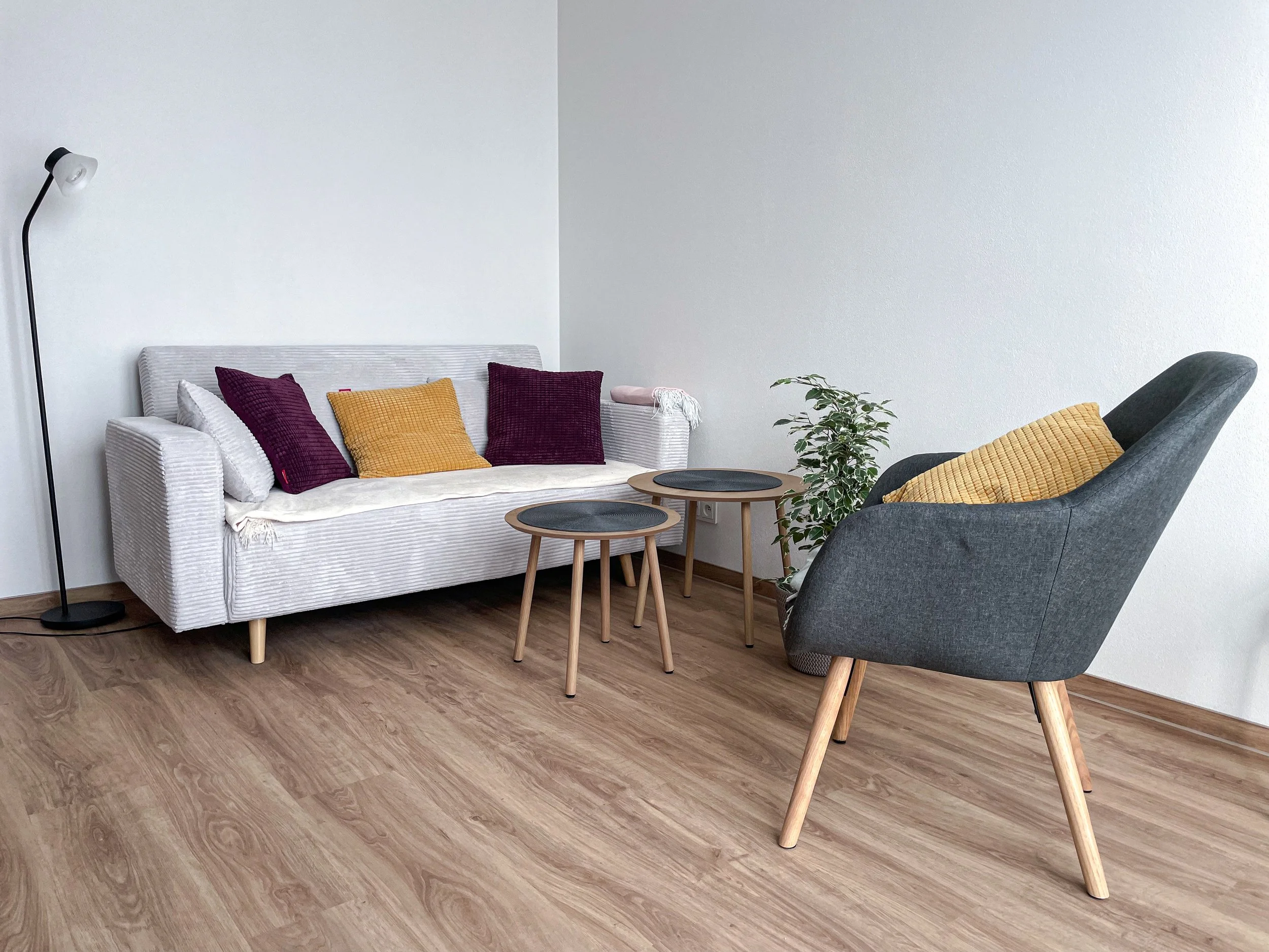 Living room with a white sofa with purple and yellow pillows, a gray armchair with a yellow pillow, two small round black-top wooden tables, a plant, and a floor lamp, with wooden flooring and plain white walls.