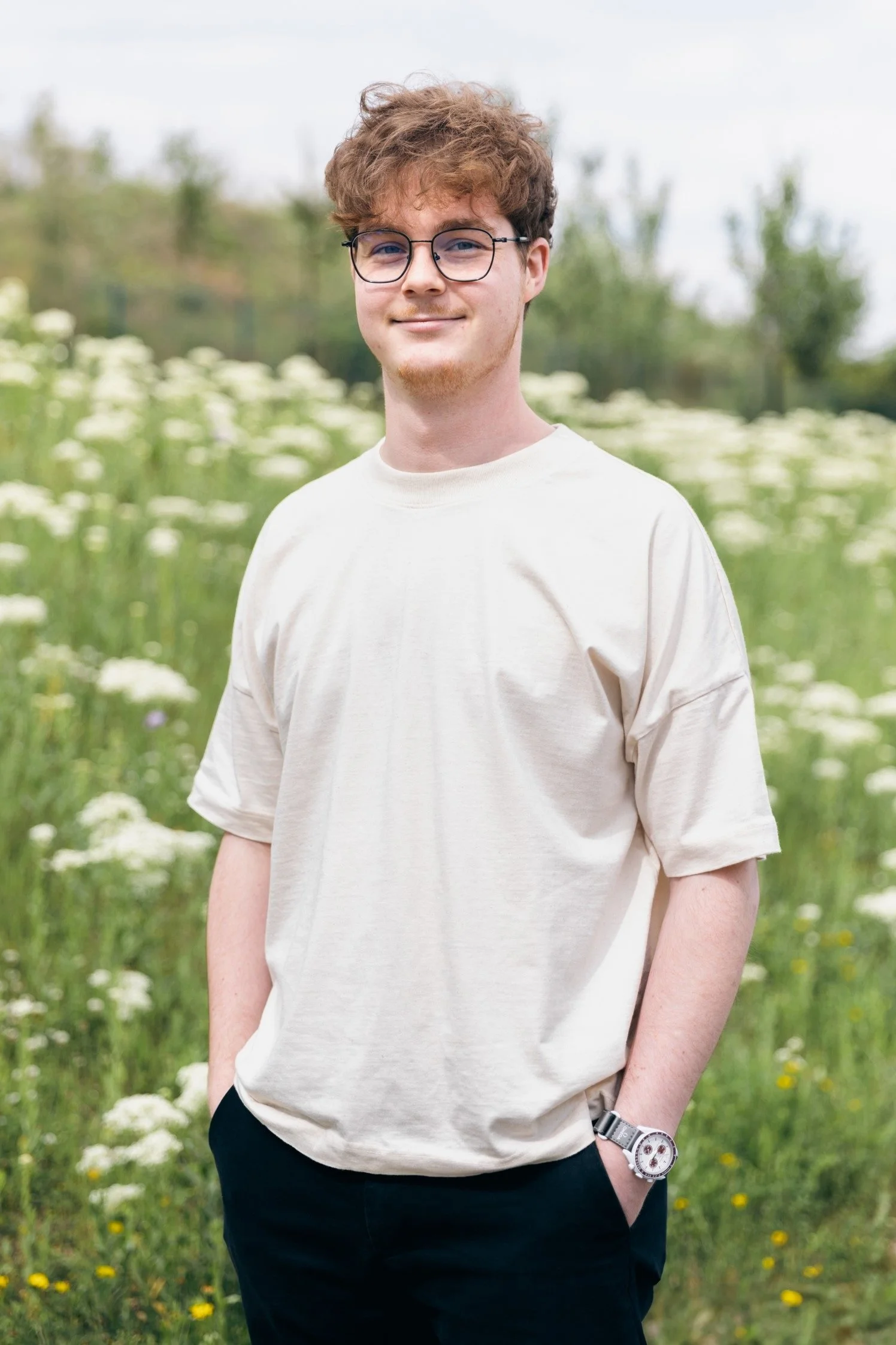 A young man with curly brown hair and glasses standing outdoors in a field of white flowers, wearing a cream-colored t-shirt and a watch on his left wrist.
