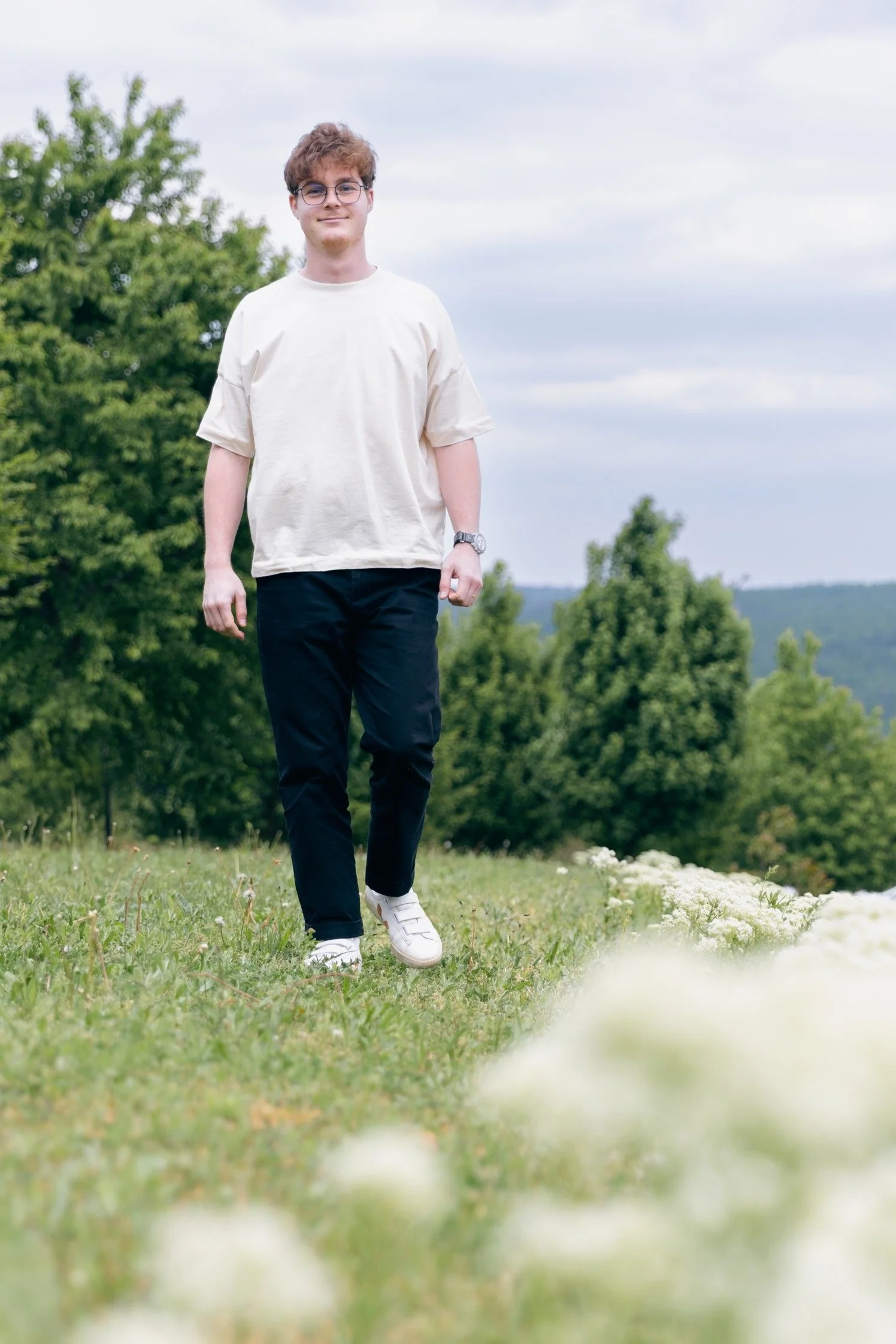 A young man with curly brown hair, glasses, wearing a beige t-shirt, black pants, and white sneakers walking outdoors in a grassy field with trees in the background.