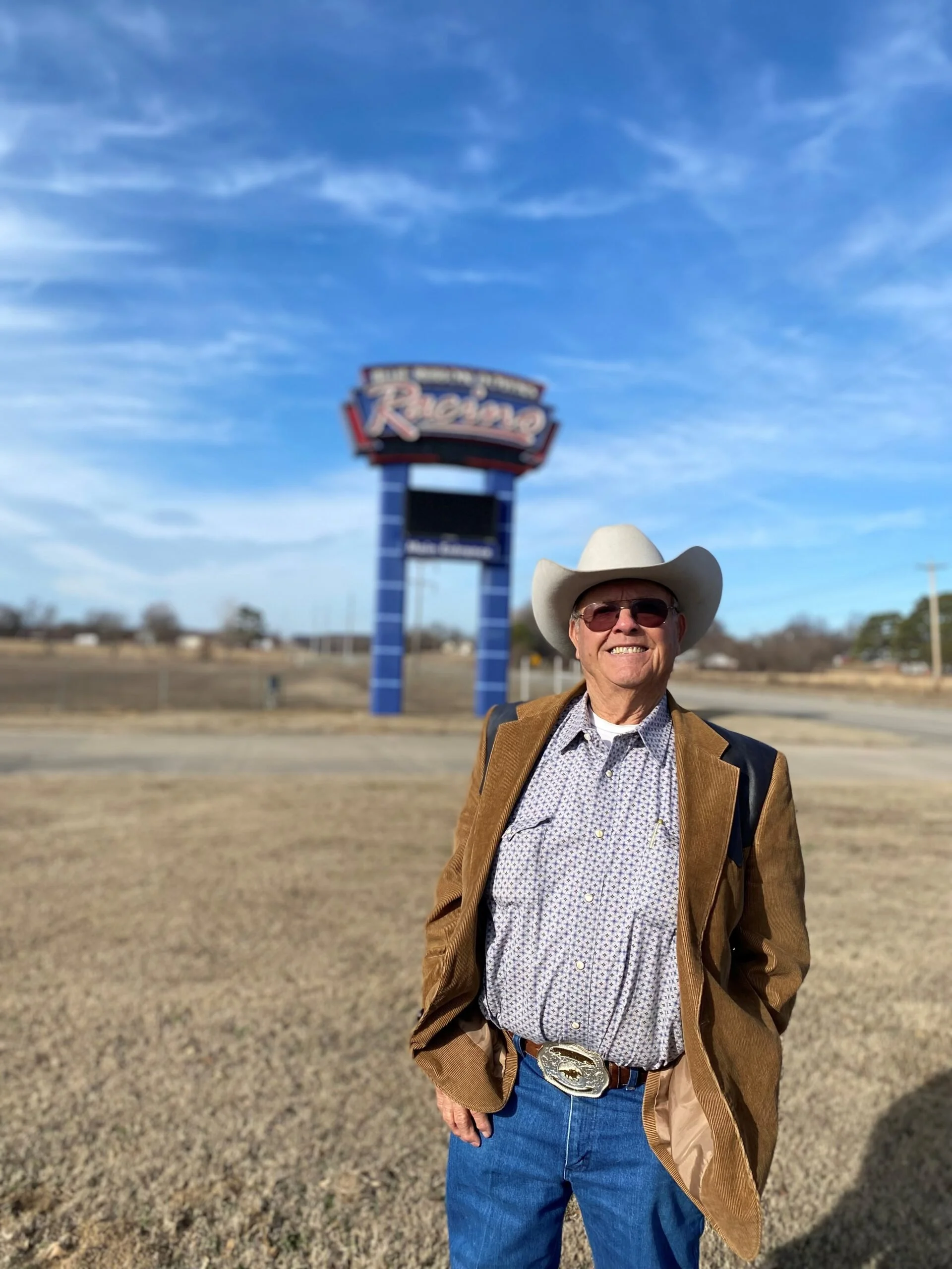A man wearing a cowboy hat, sunglasses, a patterned shirt, a brown blazer, and jeans stands outdoors in front of a blurred sign that reads 'Ranch' against a blue sky with some clouds.