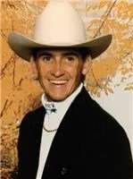 Man wearing a white cowboy hat, black suit, and white shirt, smiling in front of autumn trees.