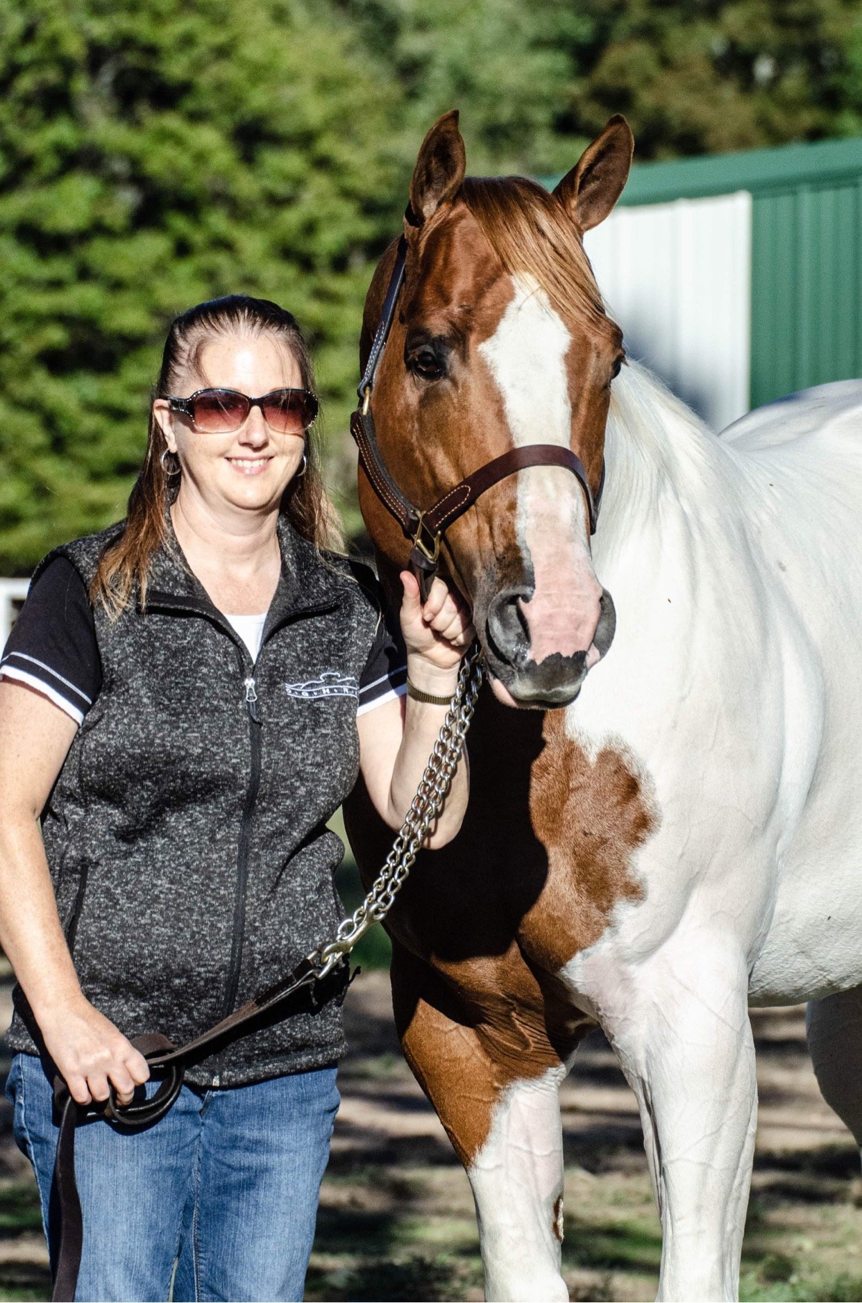 Woman standing next to a large paint horse, holding the horse's lead rope, outdoors with green trees and a building in the background.