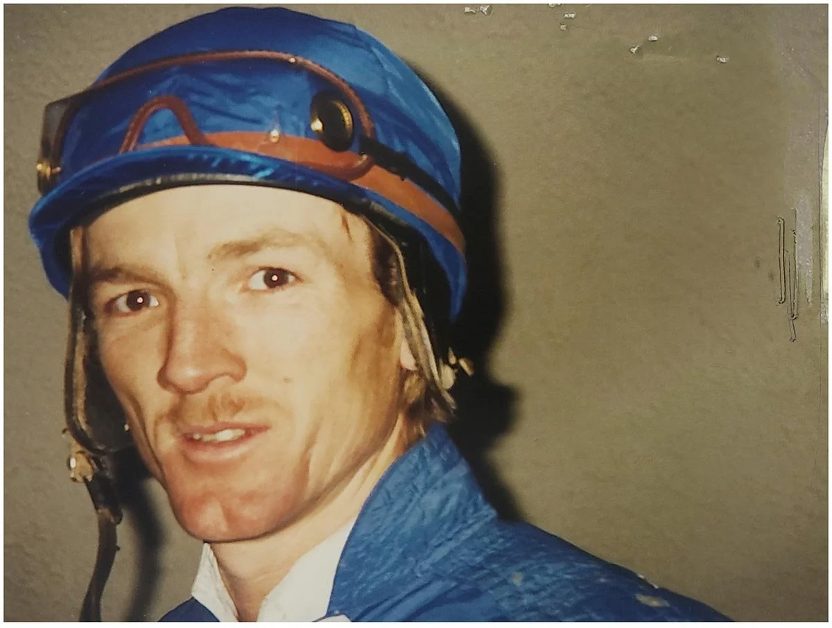 A man wearing a blue safety helmet and a blue jacket, standing indoors with a neutral wall behind him.