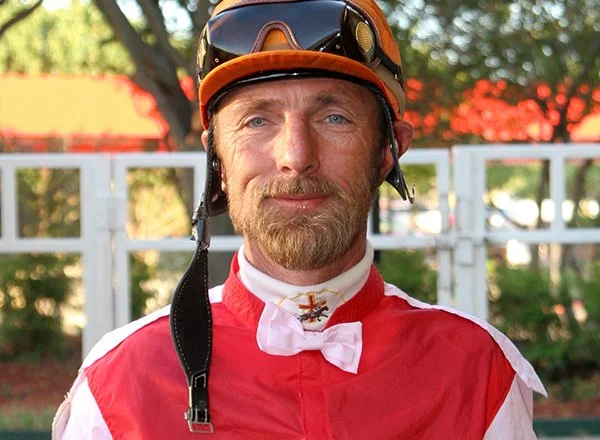 A male jockey wearing a red and white racing uniform, an orange helmet, and riding goggles, outdoors with trees in the background.