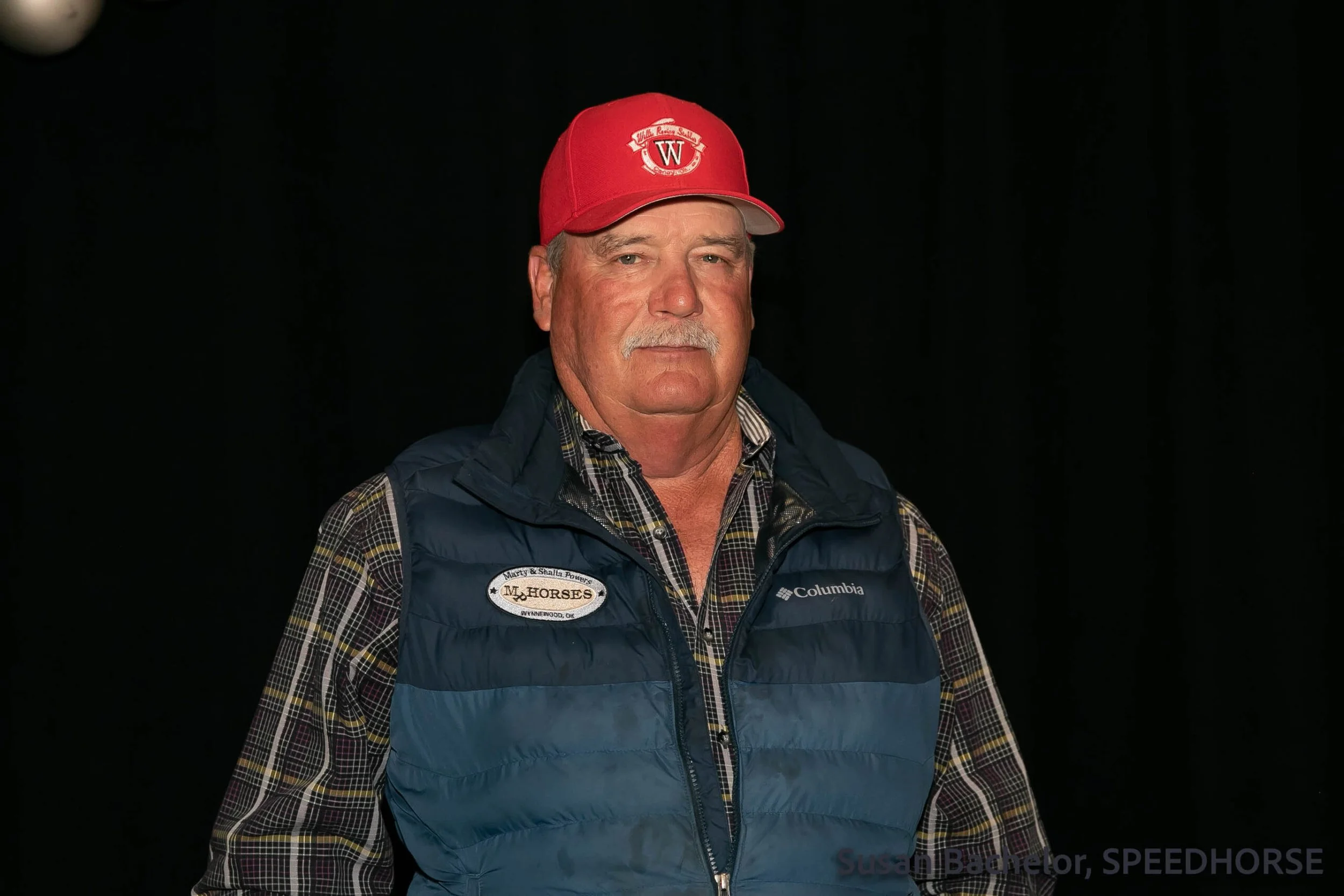 A middle-aged man wearing a red baseball cap, a blue Columbia puffer vest, and a plaid shirt, standing against a black background.