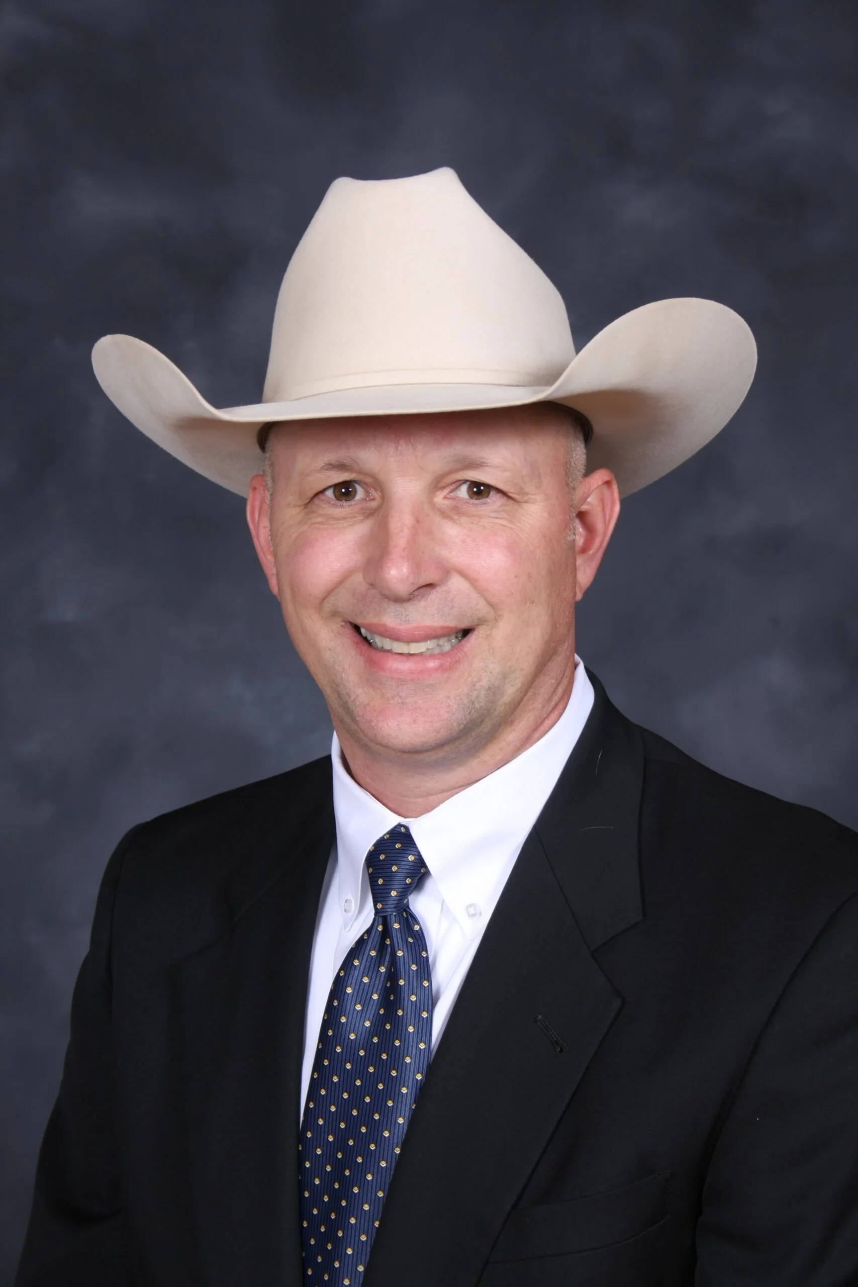 A man wearing a beige cowboy hat, a white shirt, a dark suit jacket, and a navy blue tie with yellow polka dots, smiling at the camera against a cloudy gray background.