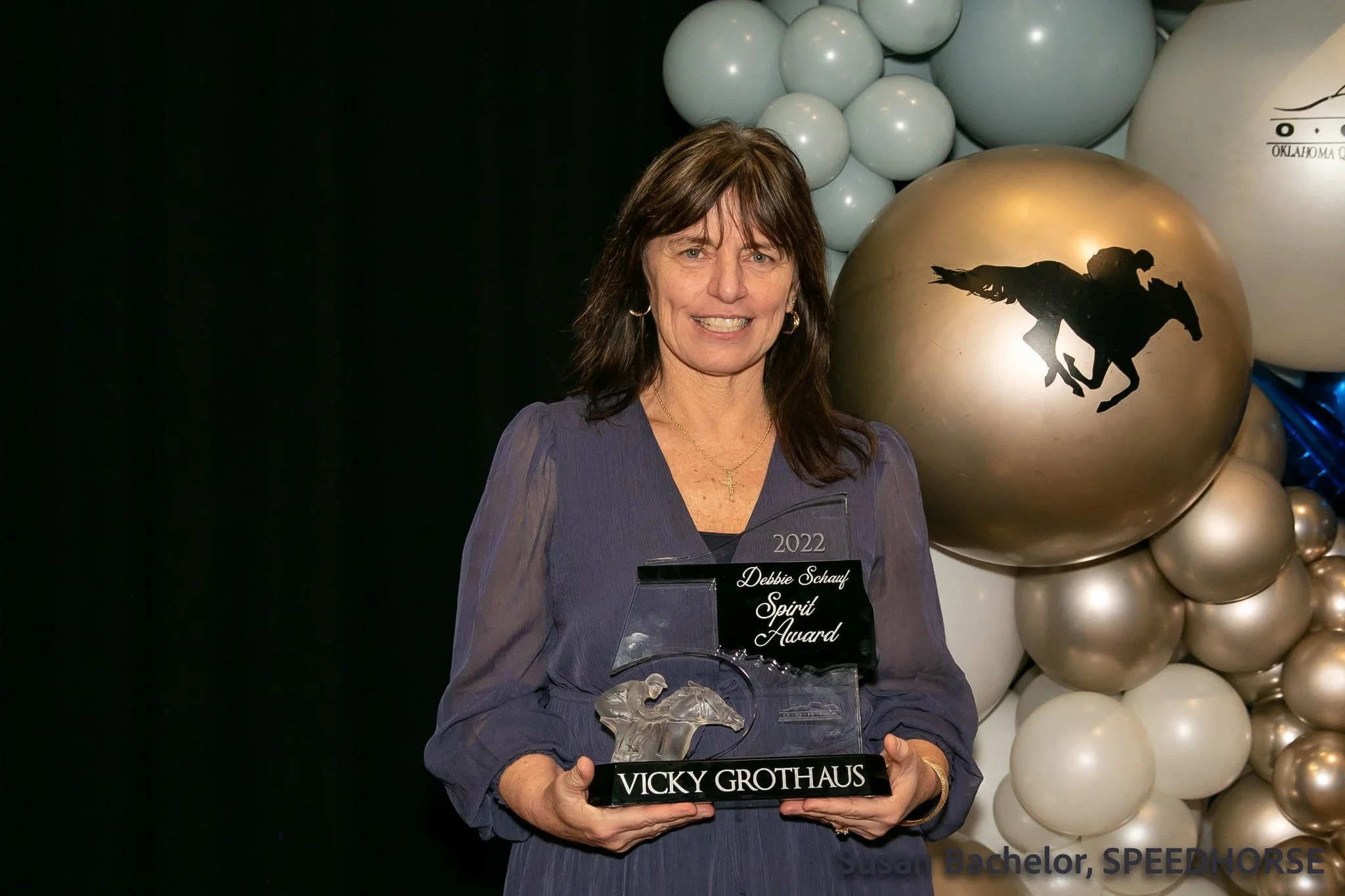 A woman smiling and holding an award plaque that reads 'Debbie Schaaf Spirit Award 2022' with the name 'Vicky Grothaus' on it. She is standing in front of a backdrop of balloons, including a large gold balloon with a silhouette of a jockey riding a horse.
