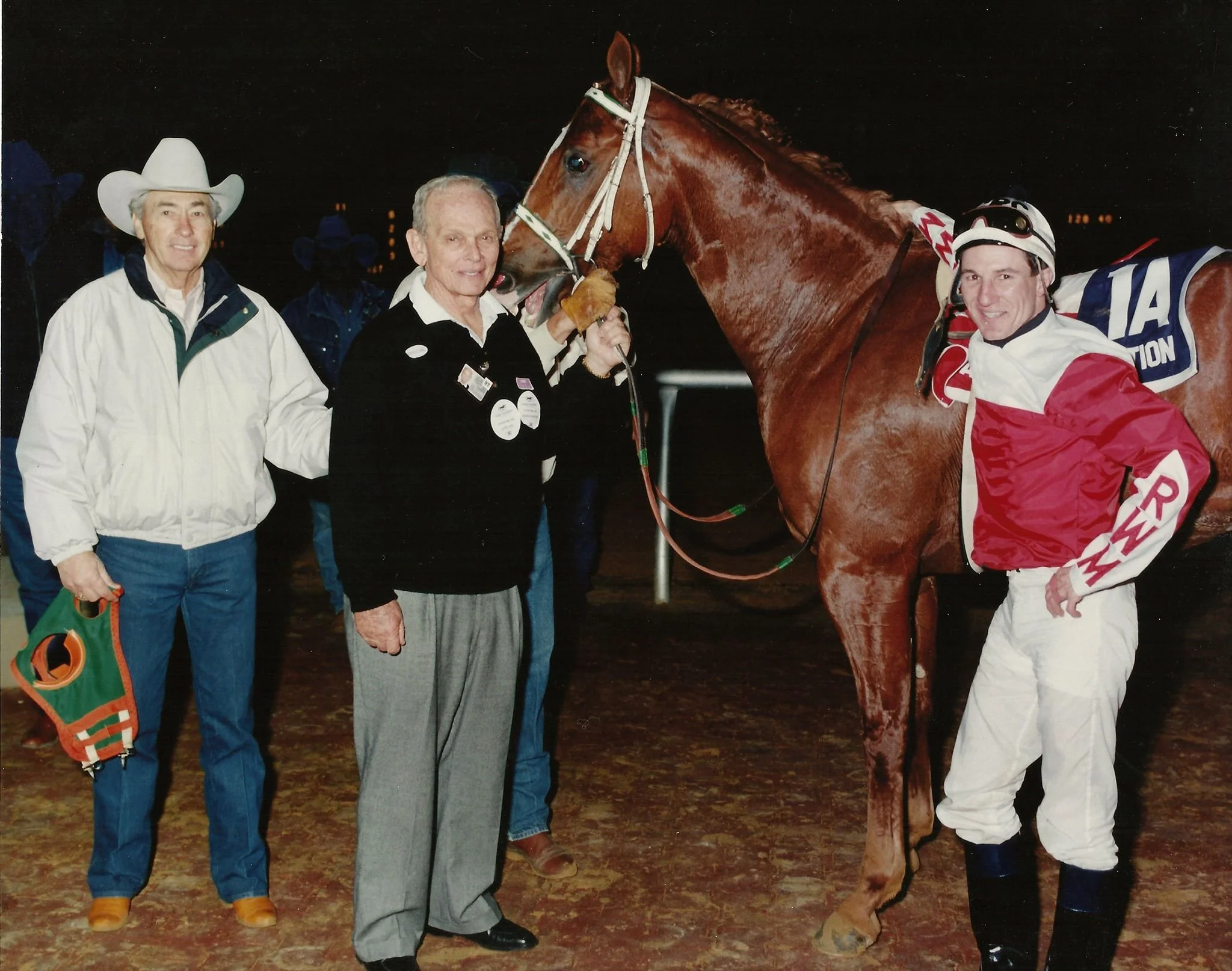 A group of three men with a winning horse at a race track at night. The man in the middle is holding the horse's bridle. The man on the right is wearing racing silks and a helmet, smiling. The man on the left is holding a bag of what appears to be prize money or awards, and all three are standing on dirt.