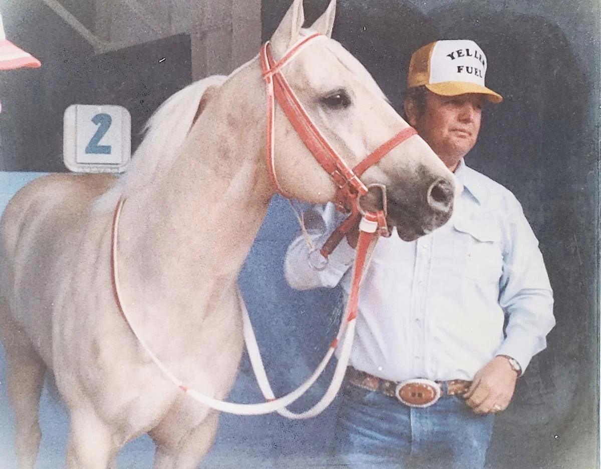 A man in a white shirt and cowboy hat standing next to a light-colored racehorse with a red halter, at a racetrack, with a blue number 2 sign in the background.