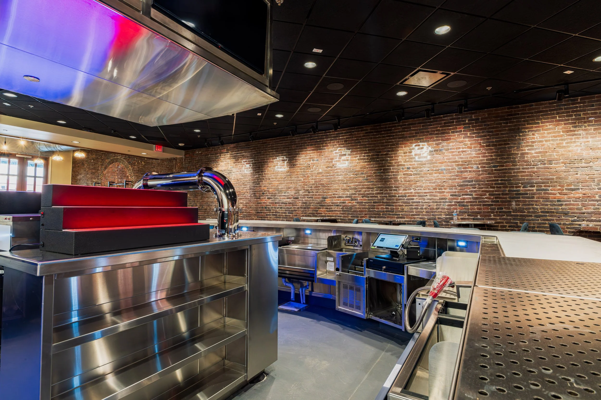 Interior of a modern restaurant kitchen with stainless steel counters, brick walls, and a black ceiling with recessed lighting.