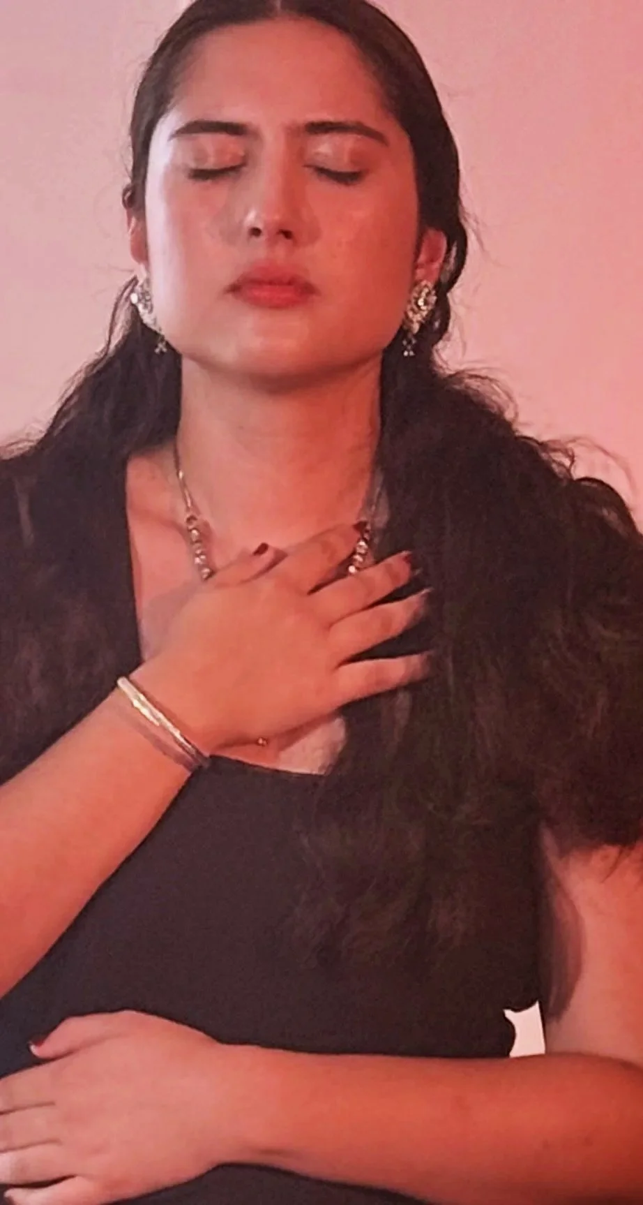 A woman with long dark hair and jewelry, eyes closed, wearing a black top, with her hand on her chest and another resting on a surface.