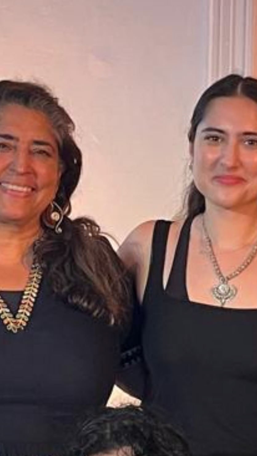 Two women standing side by side, smiling, dressed in black outfits, wearing jewelry, with a light-colored wall in the background.
