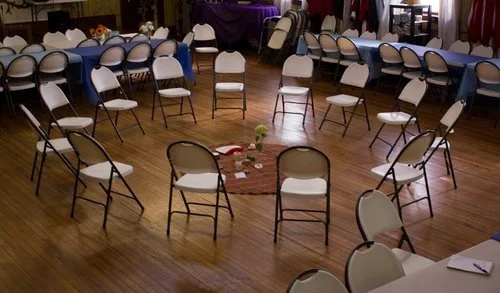 Empty banquet hall with chairs arranged in a circle and around the room, some tables with tablecloths, and scattered items on the floor.