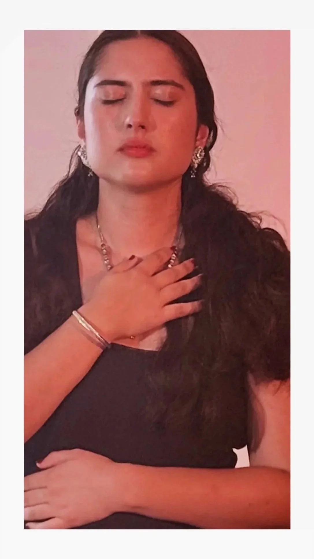 A woman with dark hair and jewelry, eyes closed, holding her hand over her chest, seated in front of a pink background.