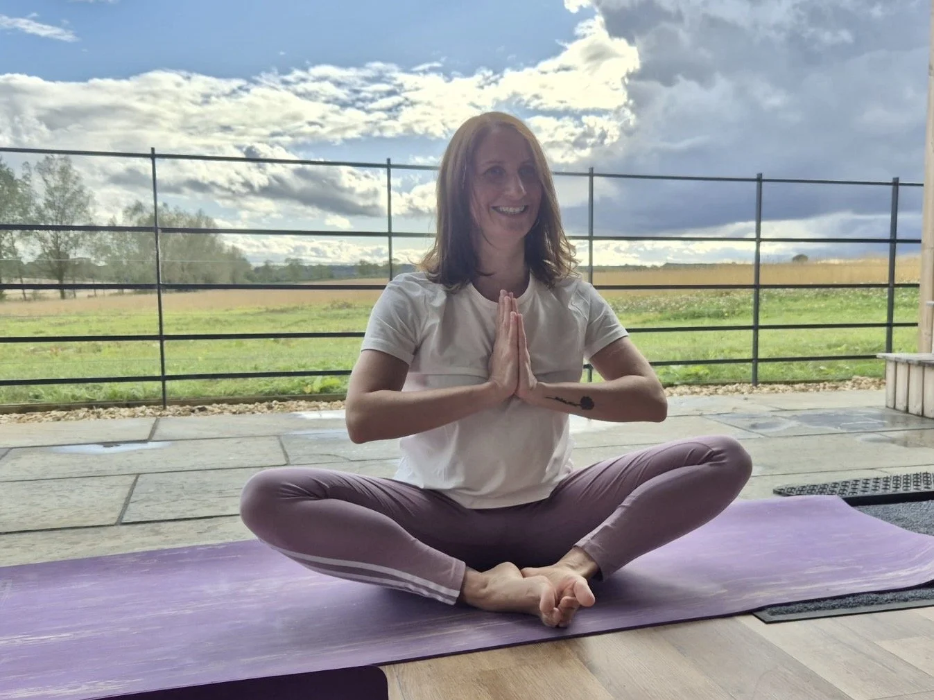 A woman practicing yoga outdoors on a purple mat, sitting cross-legged with hands in a prayer position, smiling, with a rural landscape of green fields and a partly cloudy sky in the background.