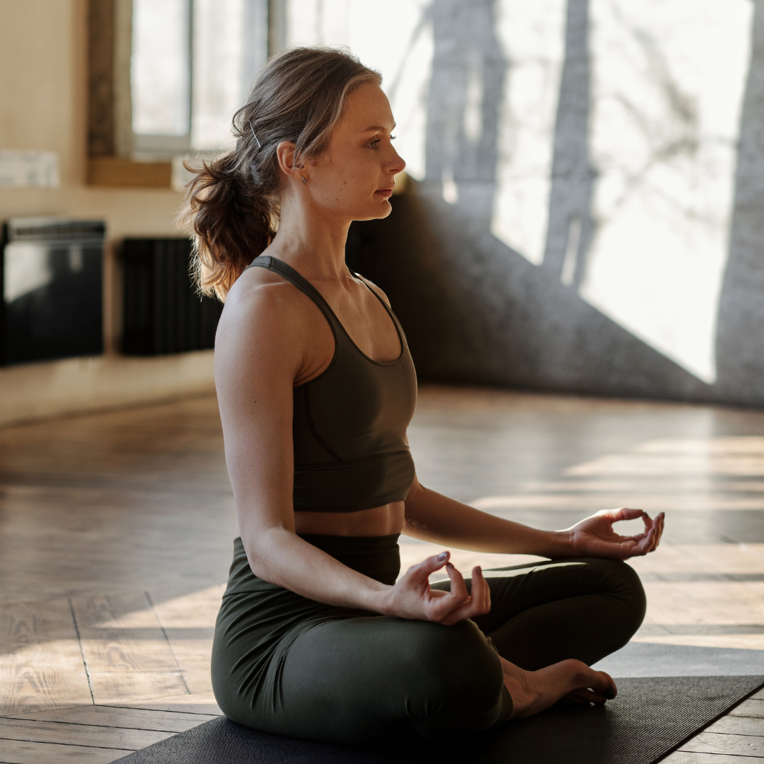 A woman practicing yoga indoors on a black mat, sitting cross-legged with hands resting on her knees, eyes closed, and sunlight streaming through windows.