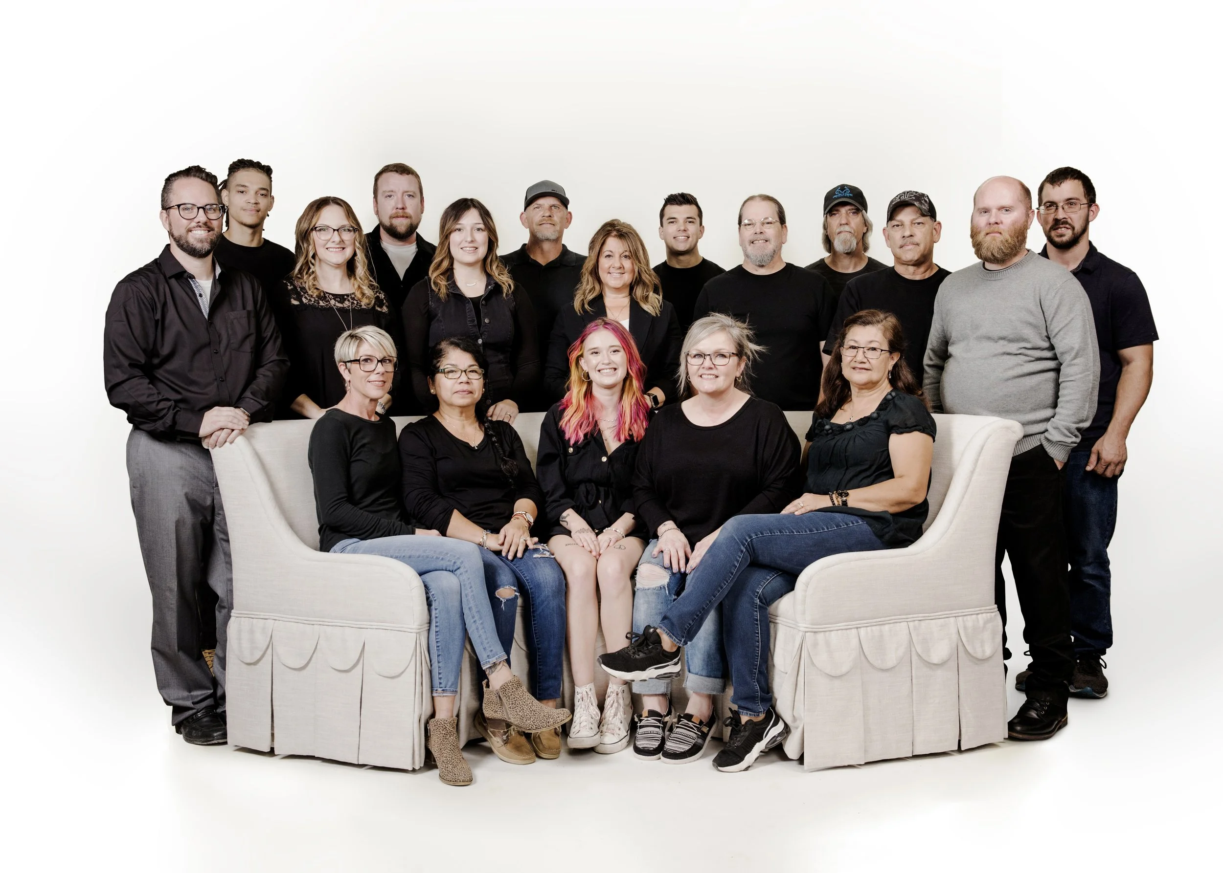 Group of 16 people posing together in a studio, some sitting on a cream-colored sofa and others standing behind it, against a plain white background.