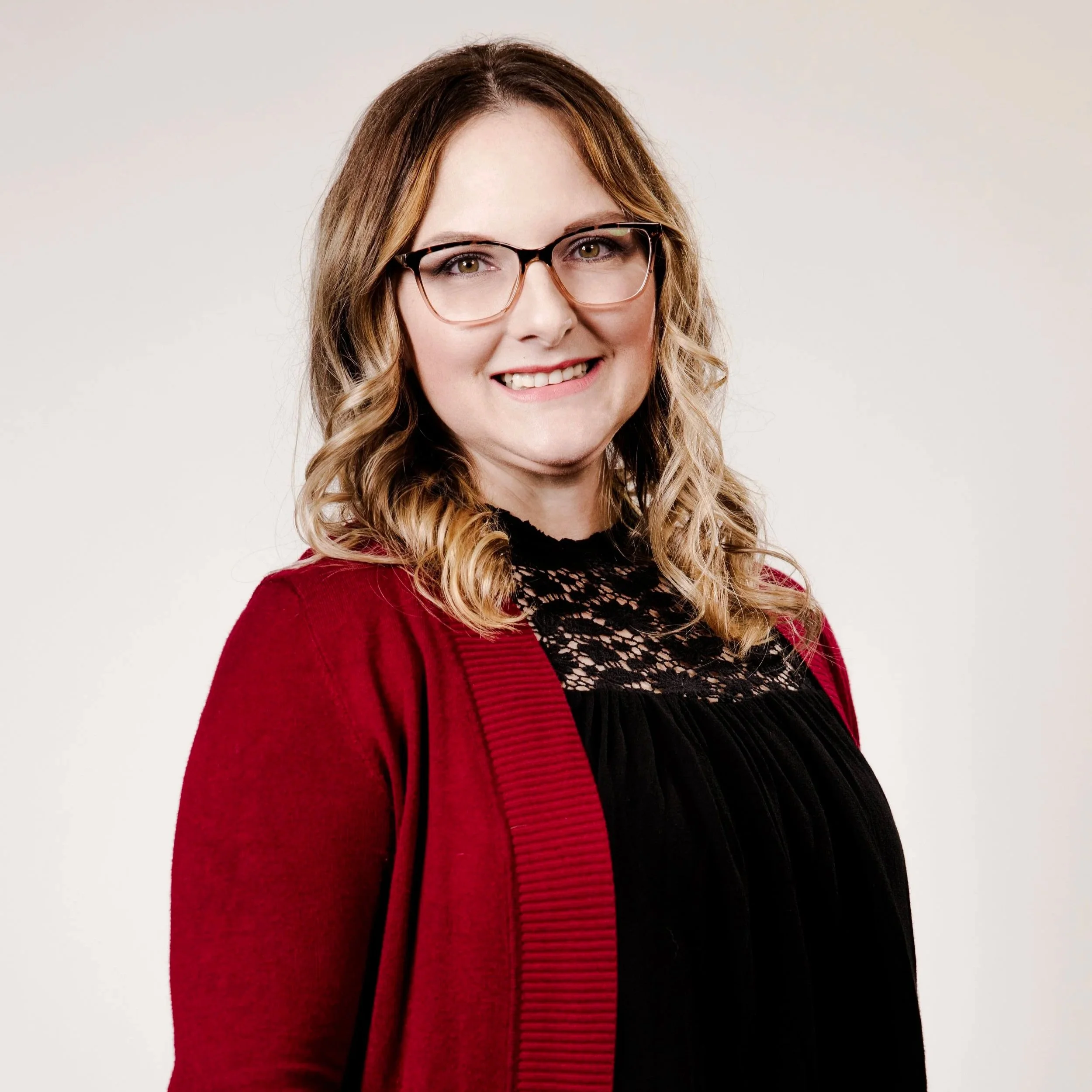 A woman with long, wavy light brown hair, wearing glasses, a black lace top, and a red cardigan, smiling at the camera against a plain white background.