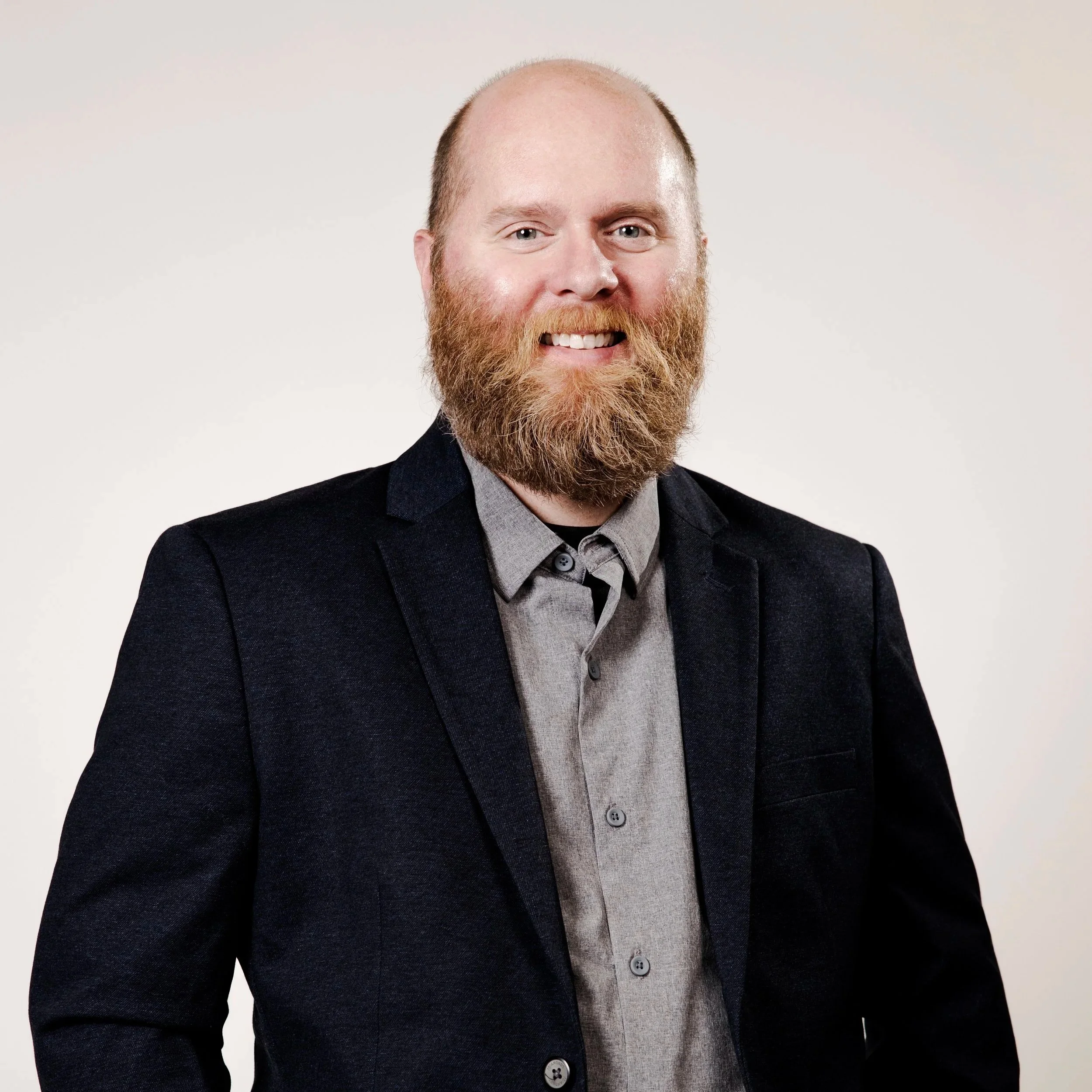 A man with a beard, smiling, wearing a black blazer and gray shirt against a plain background.