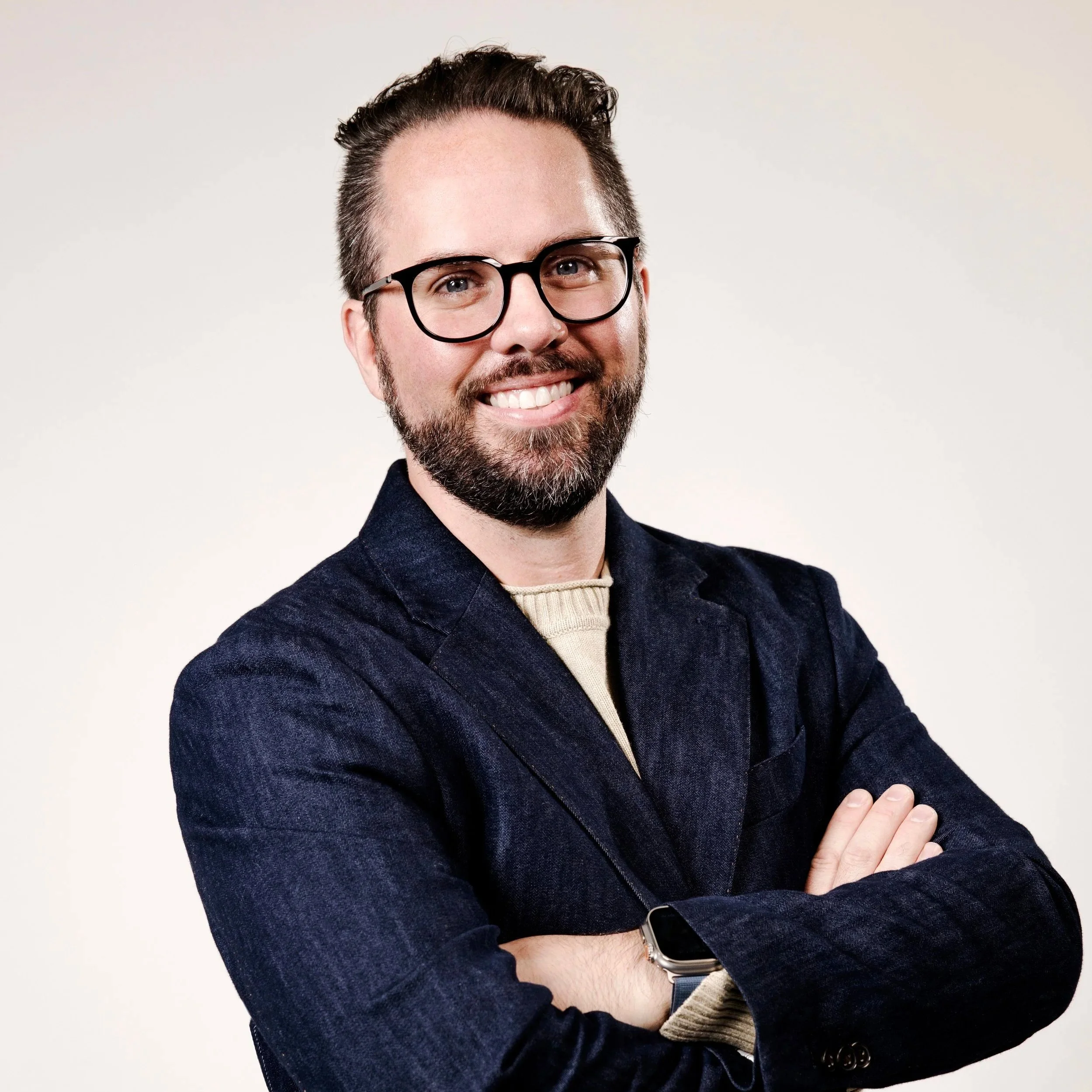 A man with glasses and a beard smiling with arms crossed, wearing a dark blazer and a beige sweater underneath, posing against a plain background.