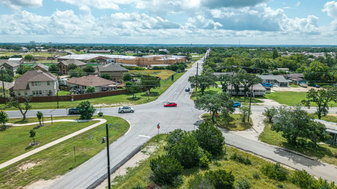 Aerial view of a suburban intersection with several cars, houses, green lawns, trees, and cloudy sky in the background on a sunny day.