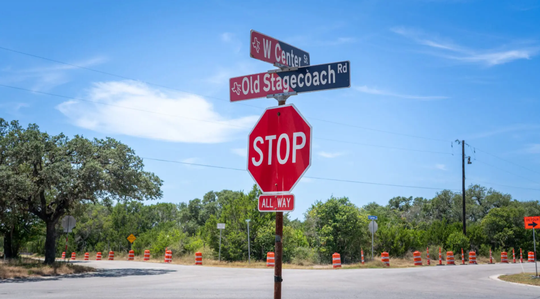 A stop sign stands at the intersection of Old Stagecoach Rd and W Center St, with orange traffic barrels lining the road and trees in the background under a blue sky.