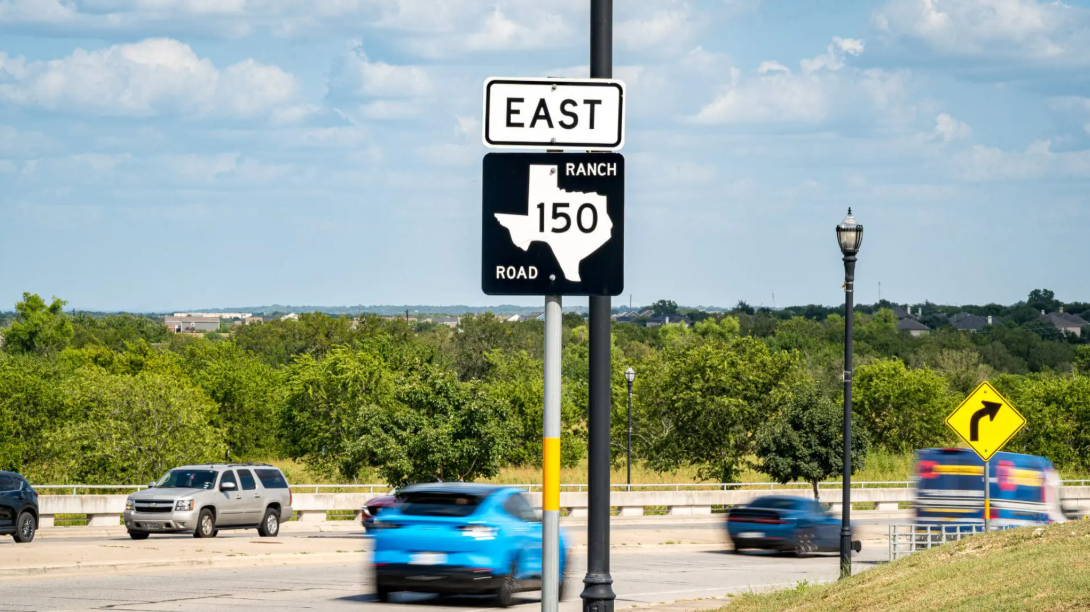 Two road signs: one reads “EAST,” the other shows a Texas state outline with “RANCH ROAD 150.” Cars drive past on a sunny day with greenery and a curve arrow sign in the background.
