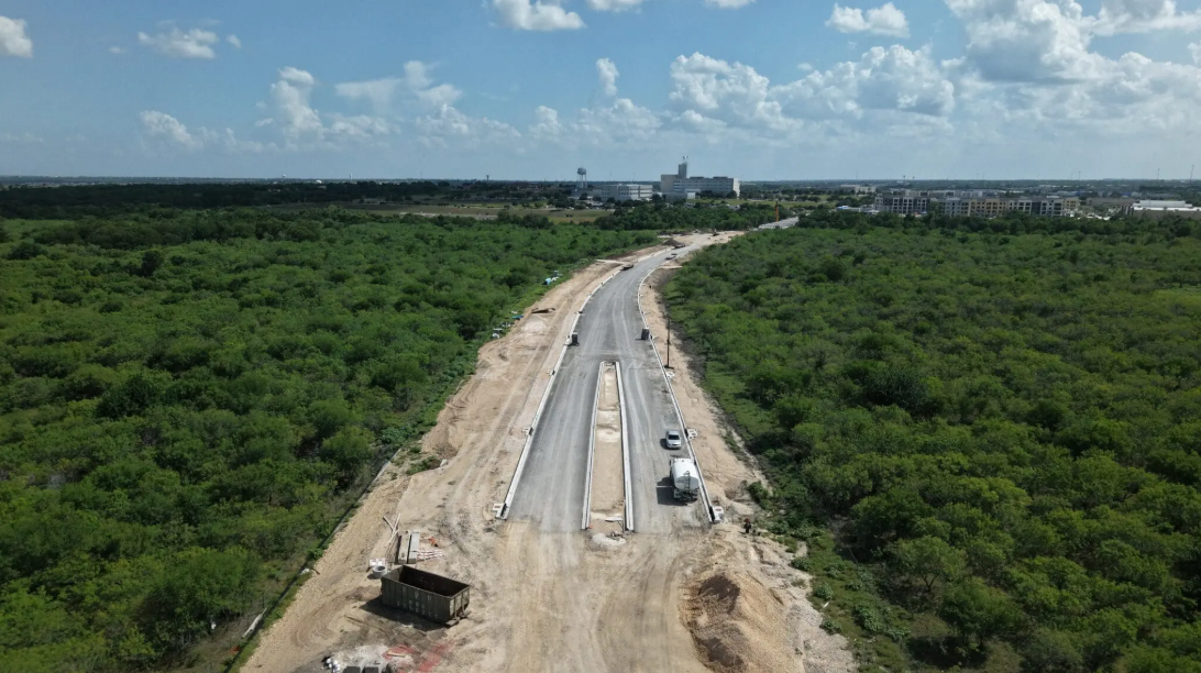 Aerial view of the partially constructed Kohlers to Seton extension cutting through dense green vegetation, with construction vehicles and equipment visible. Buildings and a water tower appear in the distant background under a partly cloudy sky in Ky