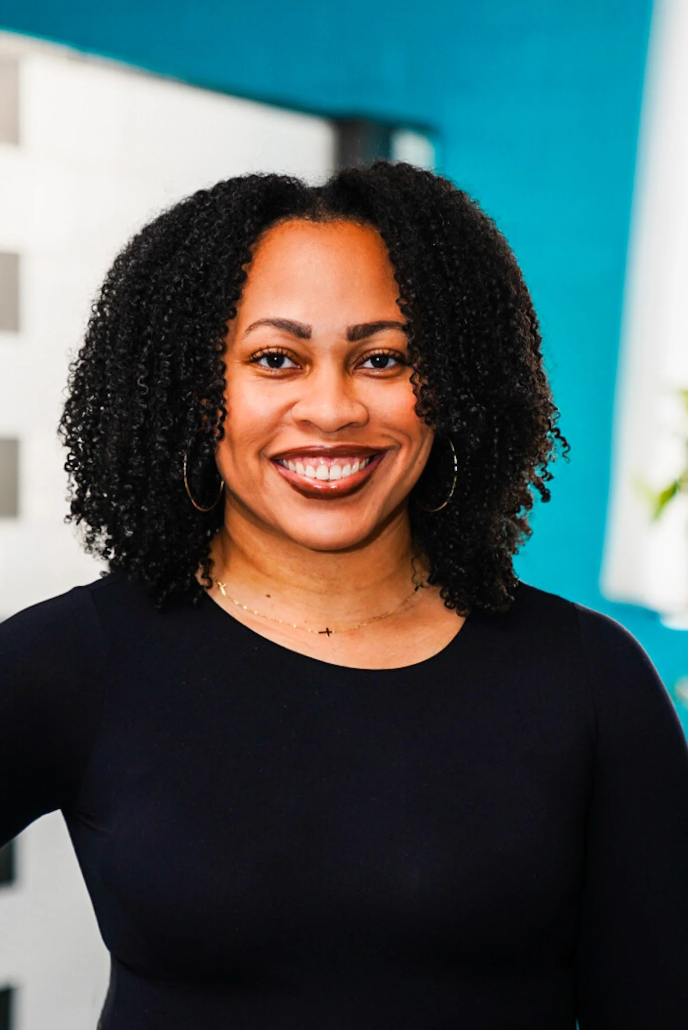 A woman with dark curly hair pulled back, smiling with arms crossed, wearing a red long-sleeve top, standing outdoors with a blurred background of trees and sunlight.