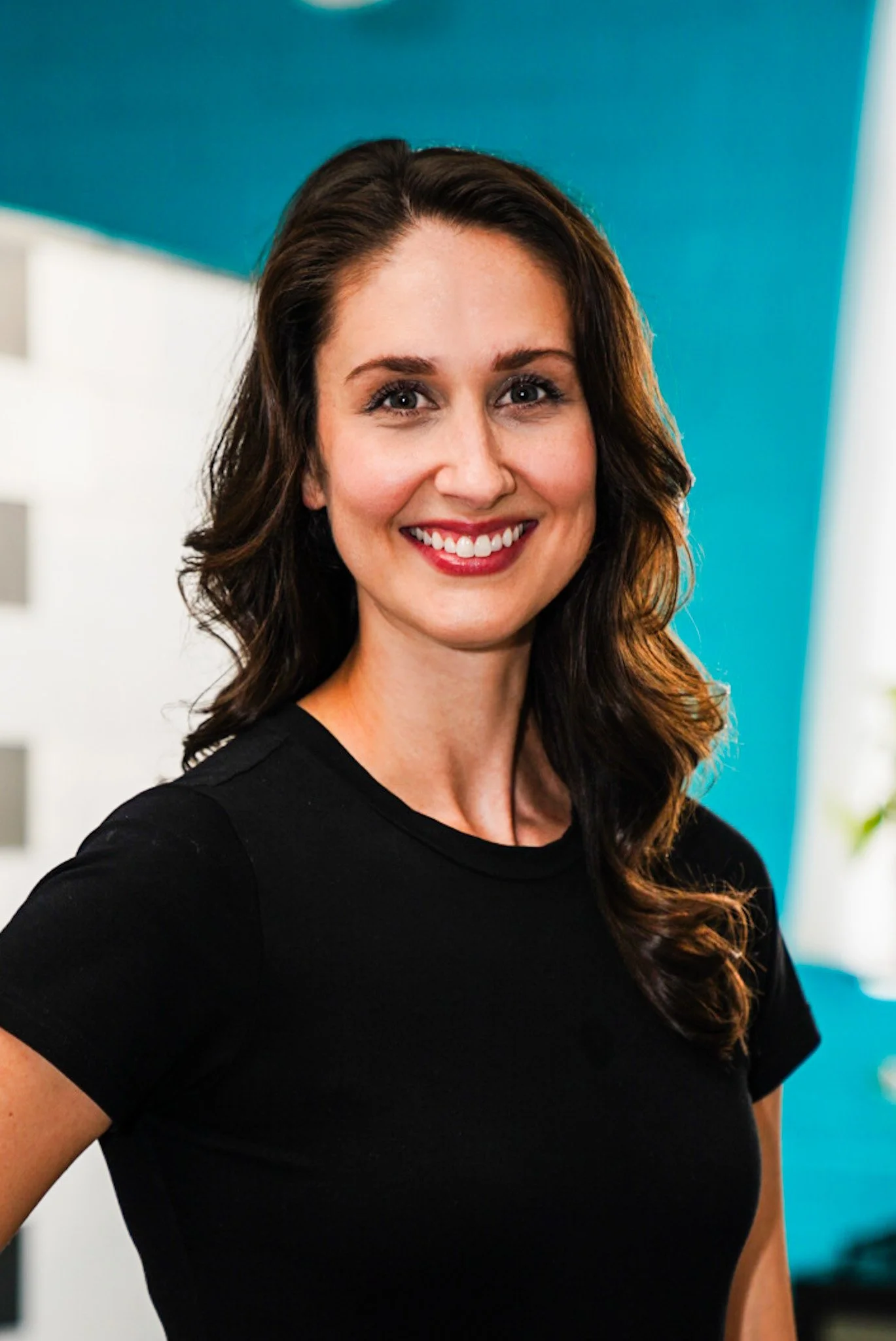 A woman with long wavy light brown hair, smiling, wearing a black top and blazer, standing against a white background.
