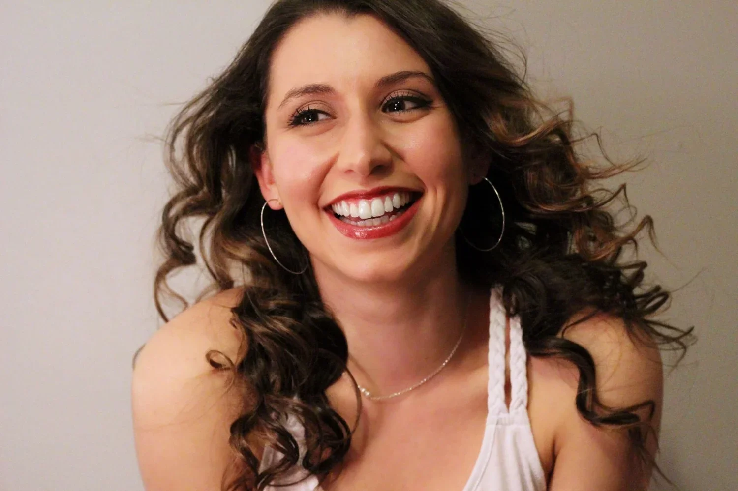 A young woman with curly brown hair smiling and showing her teeth, wearing hoop earrings and a white tank top.