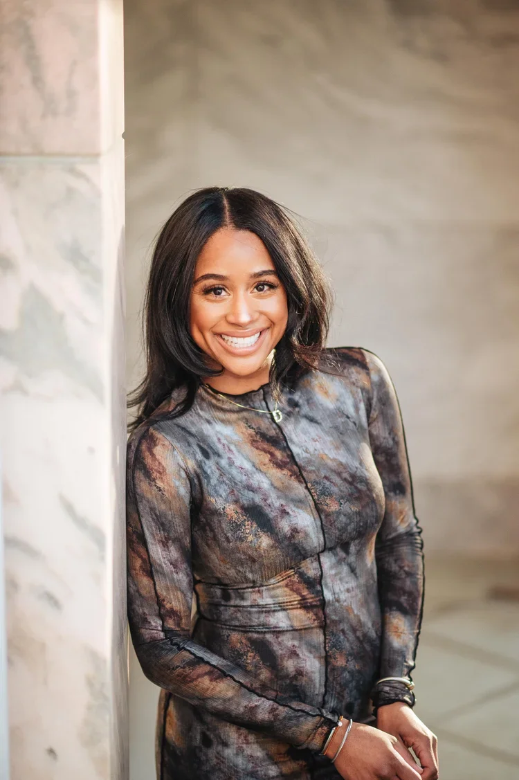 Smiling woman with shoulder-length dark hair wearing a colorful patterned long-sleeve dress, leaning against a marble wall.