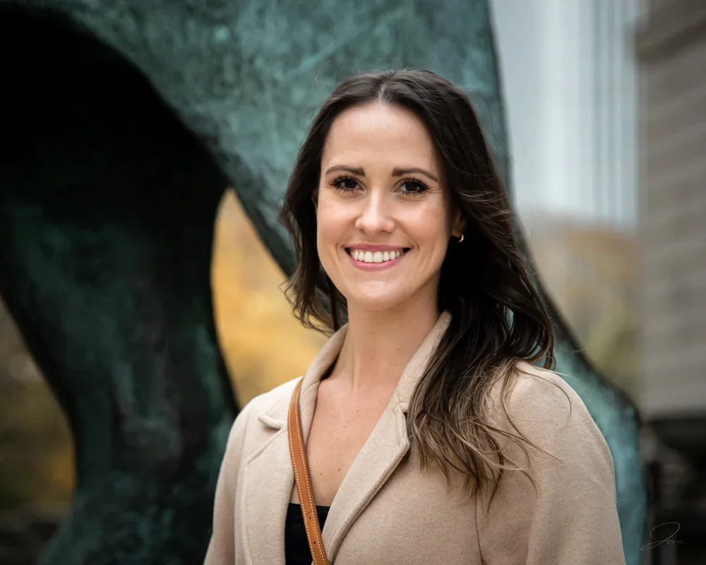 A smiling woman with long dark hair wearing a beige blazer and a black top, standing outdoors in front of a large abstract sculpture.