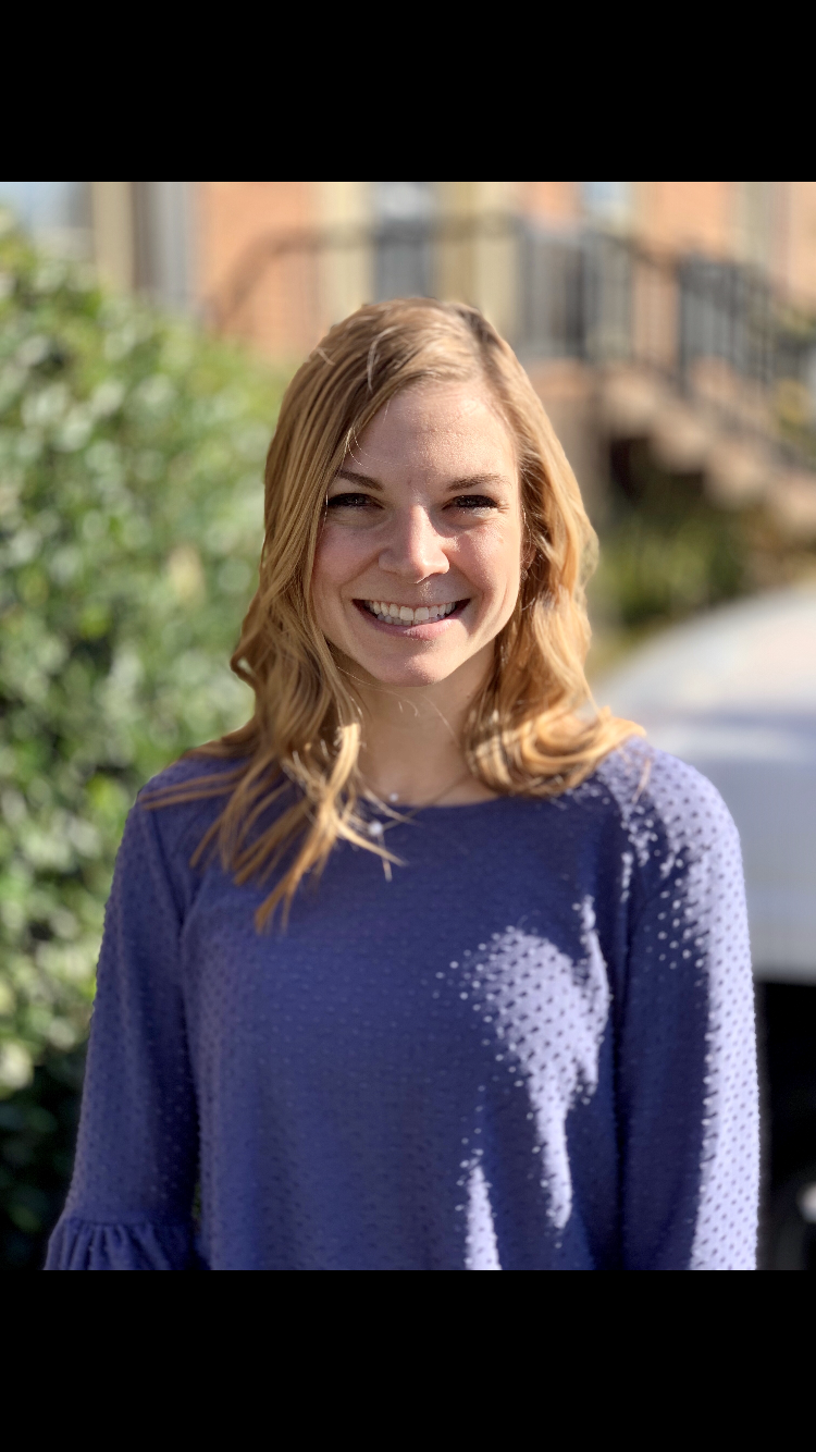 A young woman with light brown hair smiling outdoors on a sunny day, wearing a purple textured long-sleeve shirt.