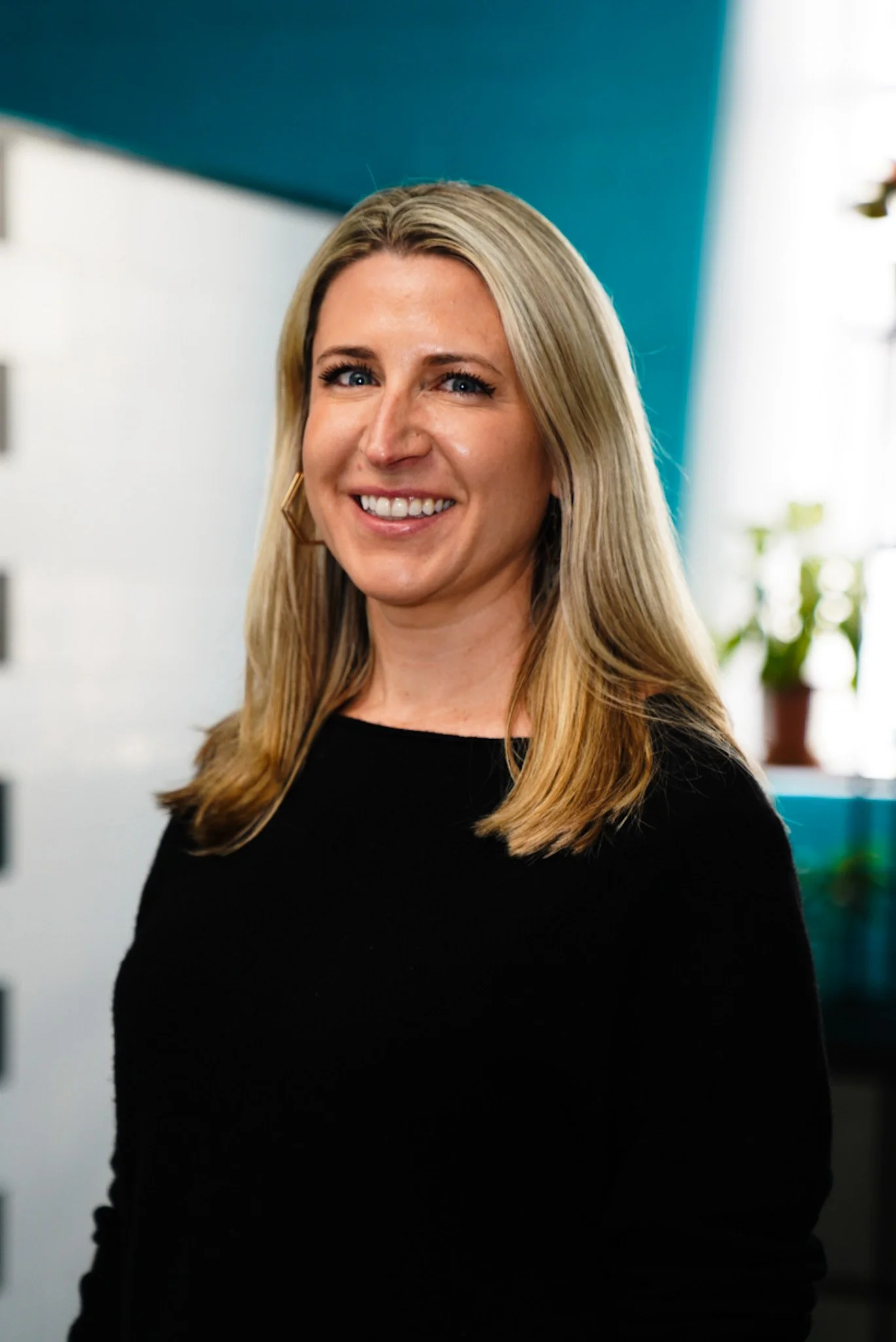 Portrait of a smiling blonde woman with blue eyes wearing a blue top and a necklace, against a gray background.