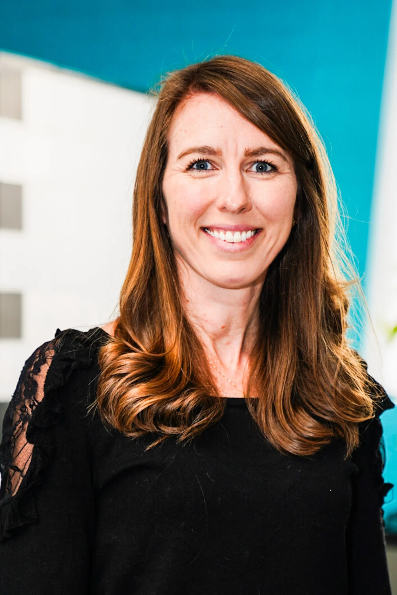 A woman with long reddish-brown hair, blue eyes, and wearing a black shirt with white lettering and yellow earrings, standing in front of a light-colored brick wall, smiling at the camera.