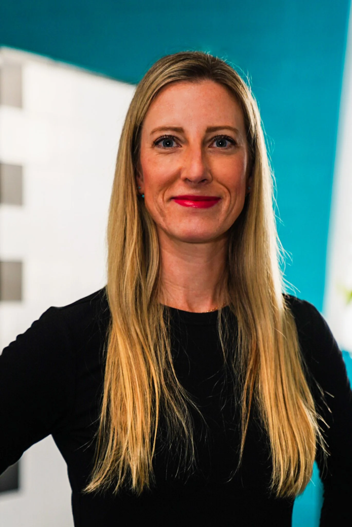 A woman with long blonde hair, green eyes, and red lipstick, wearing a blue blouse, smiling in front of a light-colored wooden background.
