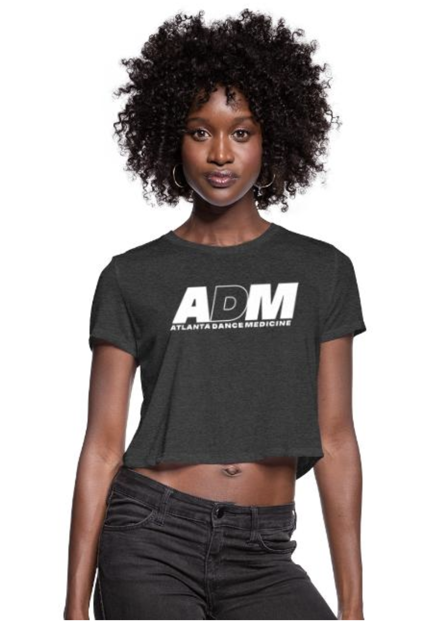 A woman with dark skin and curly hair, wearing a black crop top with the words 'Atlanta Dance Medicine' and 'ADM' on it, and black jeans.
