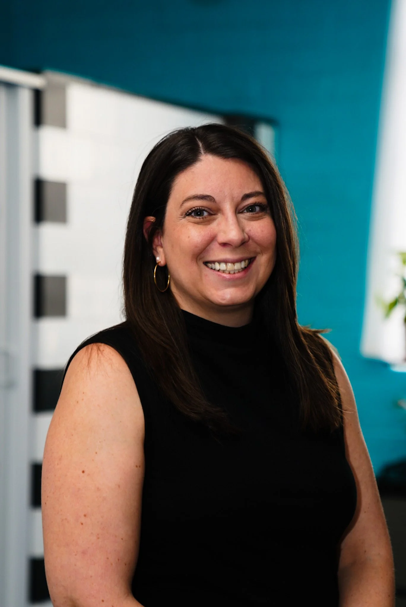 A woman with shoulder-length brown hair standing outdoors against a brick wall. She is smiling and wearing a black Atlanta Dance Medicine T-shirt.