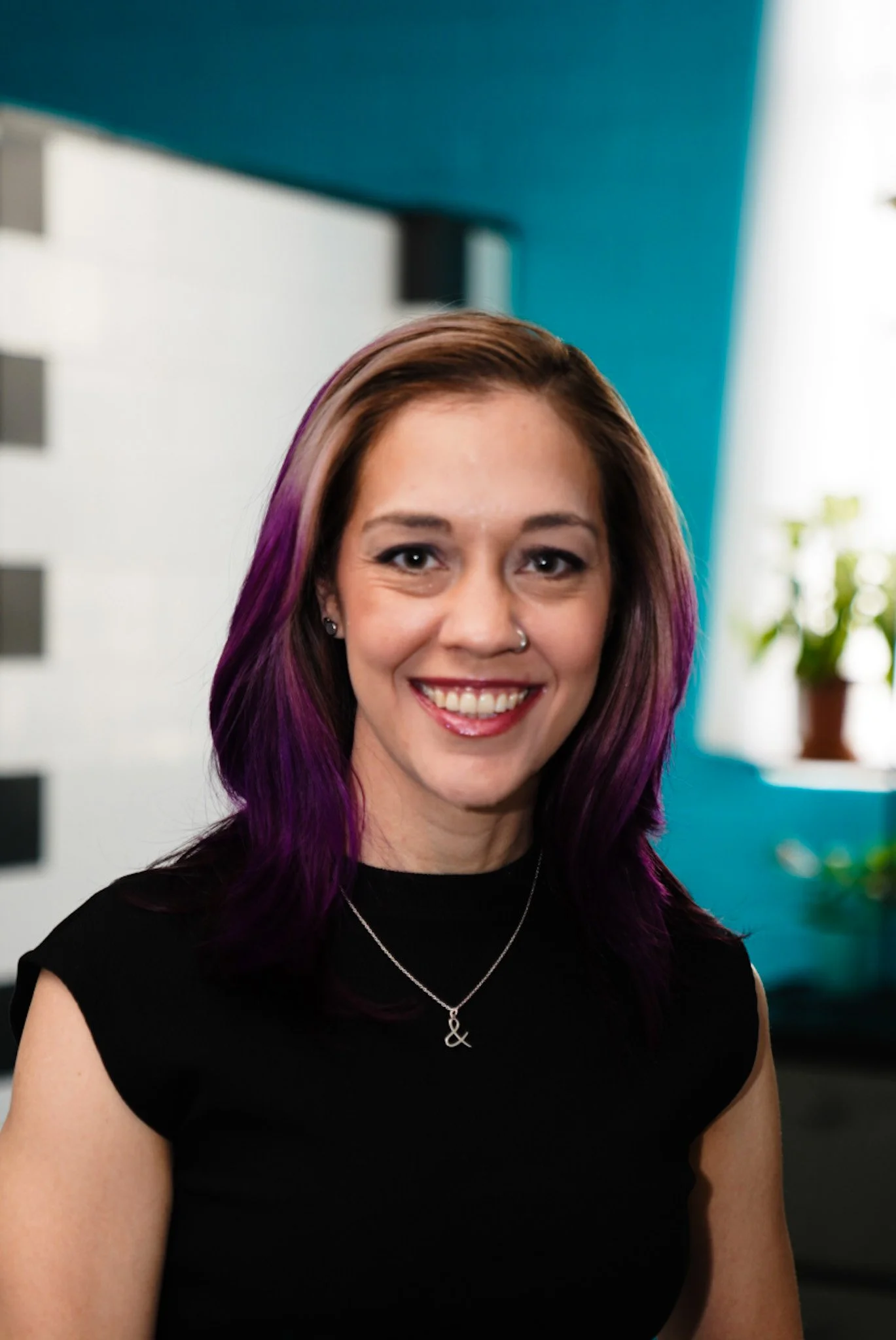 A young woman with curly blonde hair smiling outdoors in a green, wooded area.
