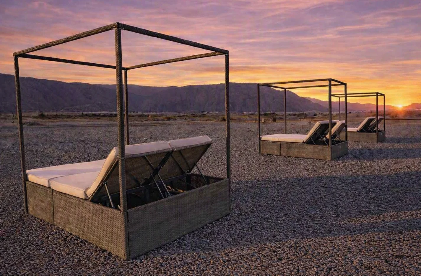 Four outdoor canopy beds with beige curtains and white cushions on a gravel surface during sunset, with mountains in the background.