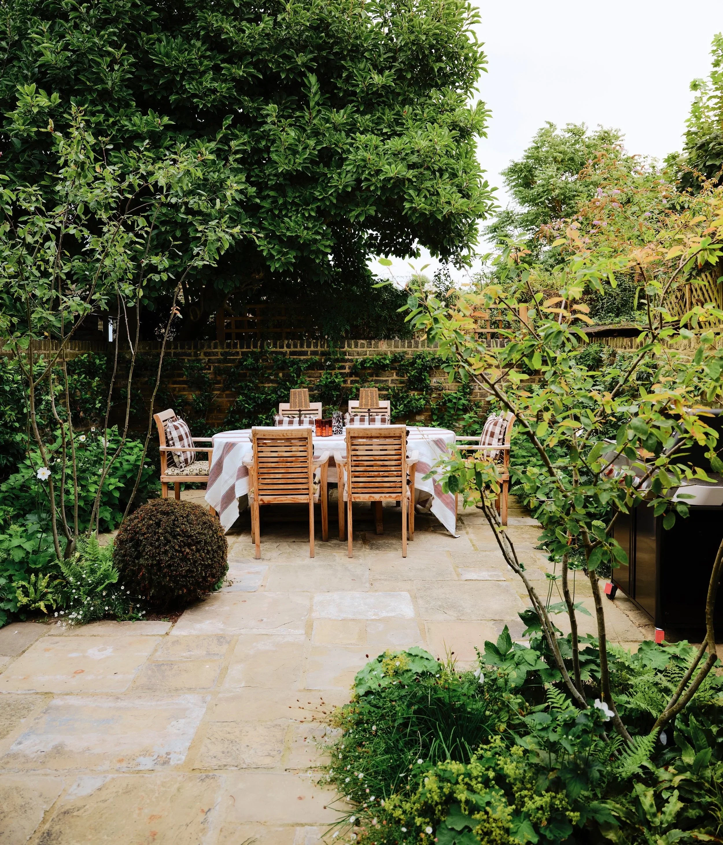 Outdoor dining area with a large rectangular table and chairs, set up in a lush garden with various green plants, trees, and a brick wall in the background.