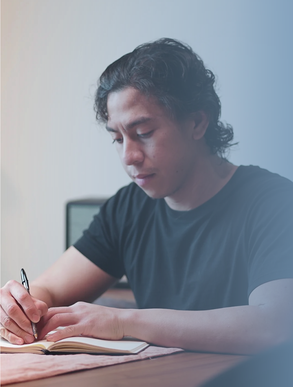 A young man with wavy dark hair, wearing a black t-shirt, sitting at a table, writing in a notebook with a pen.