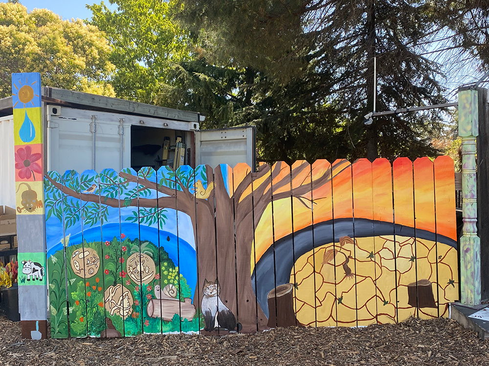 Colorful mural on a wooden fence depicting a tree with animals, a rainy cloud, and a dry cracked landscape at sunset.