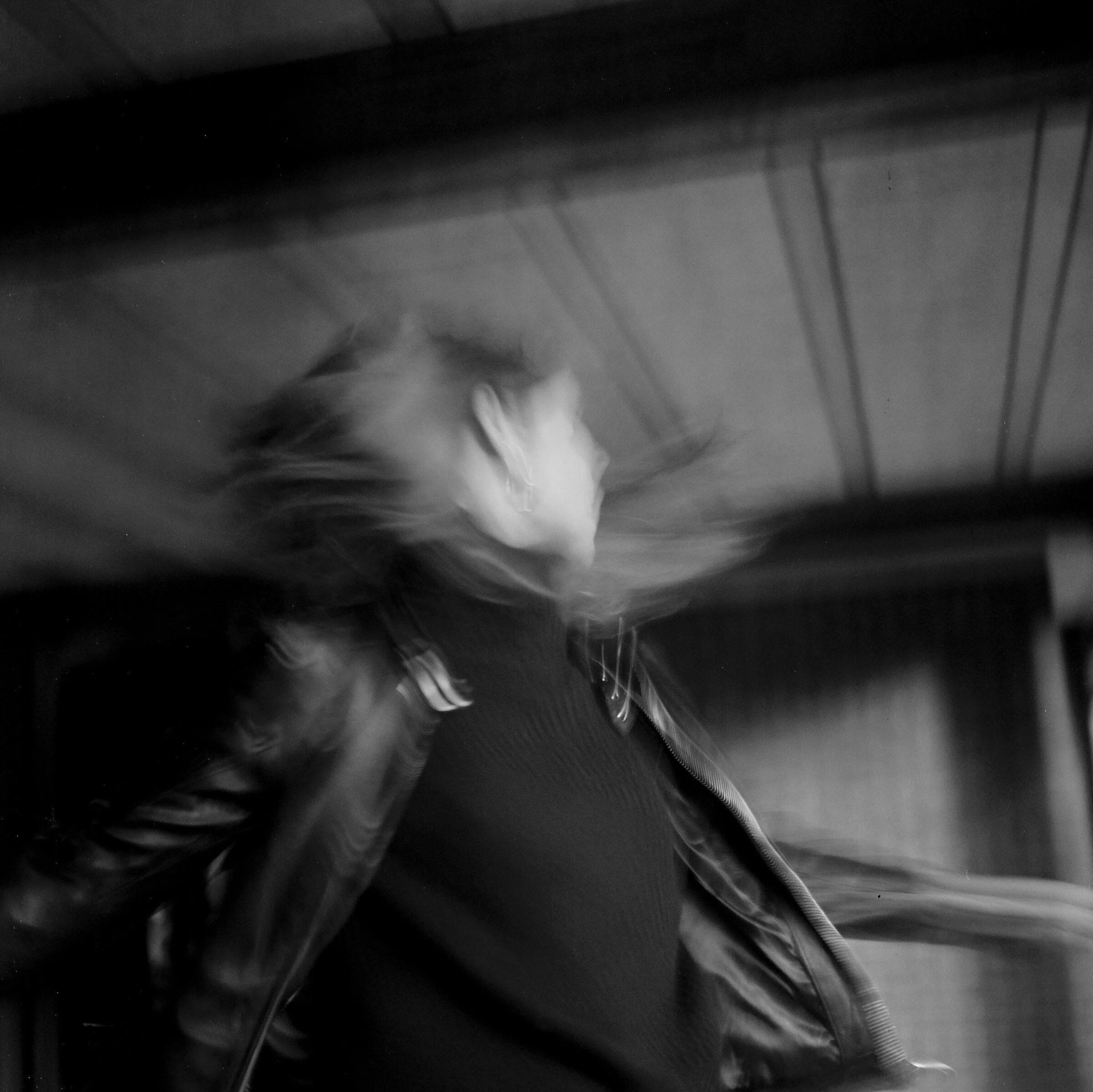 Blurred black-and-white photo of a woman with hallway background, looking to the side, wearing a jacket and sweater, with her hair in motion.