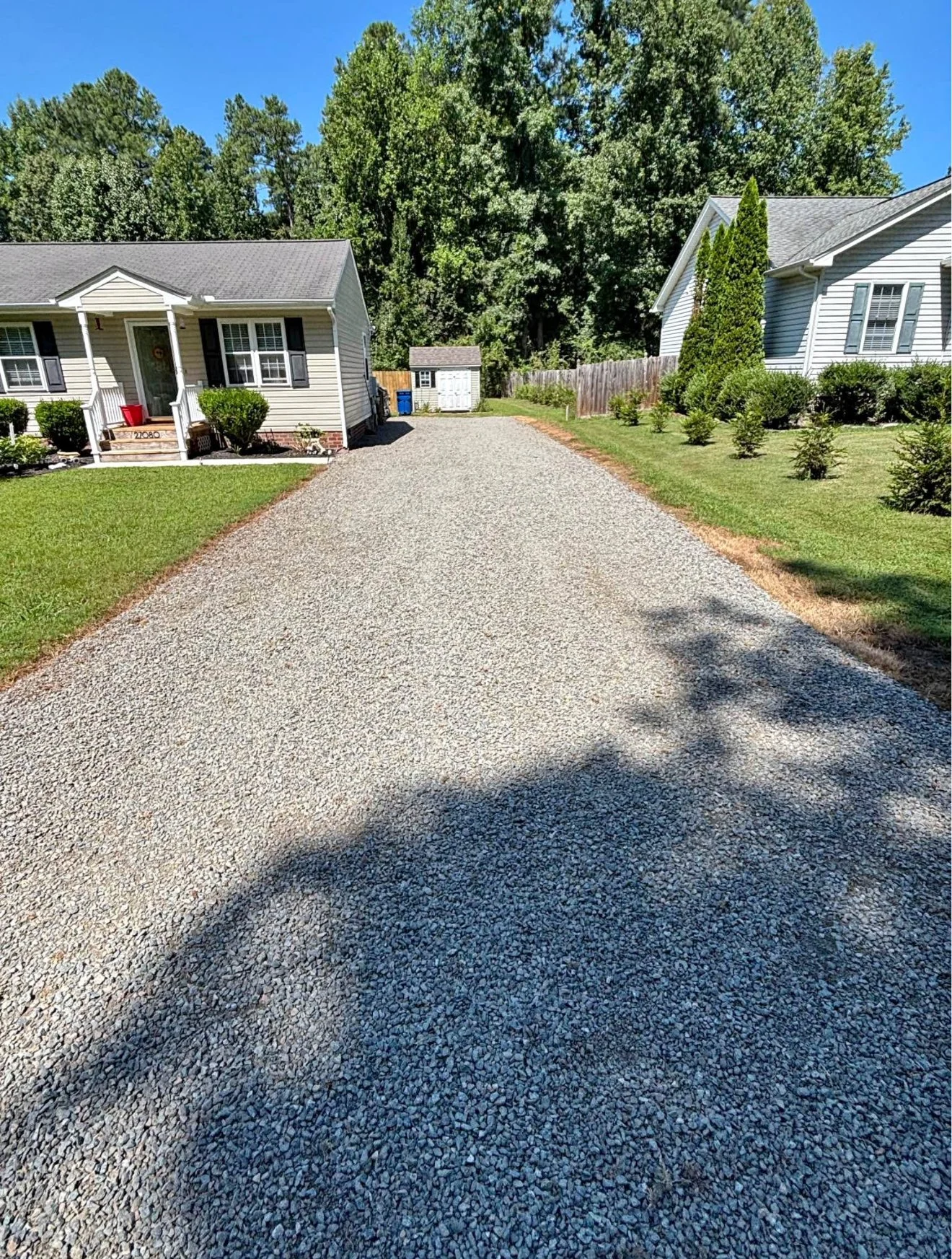 A gravel driveway between two houses with green lawns and trees in the background.