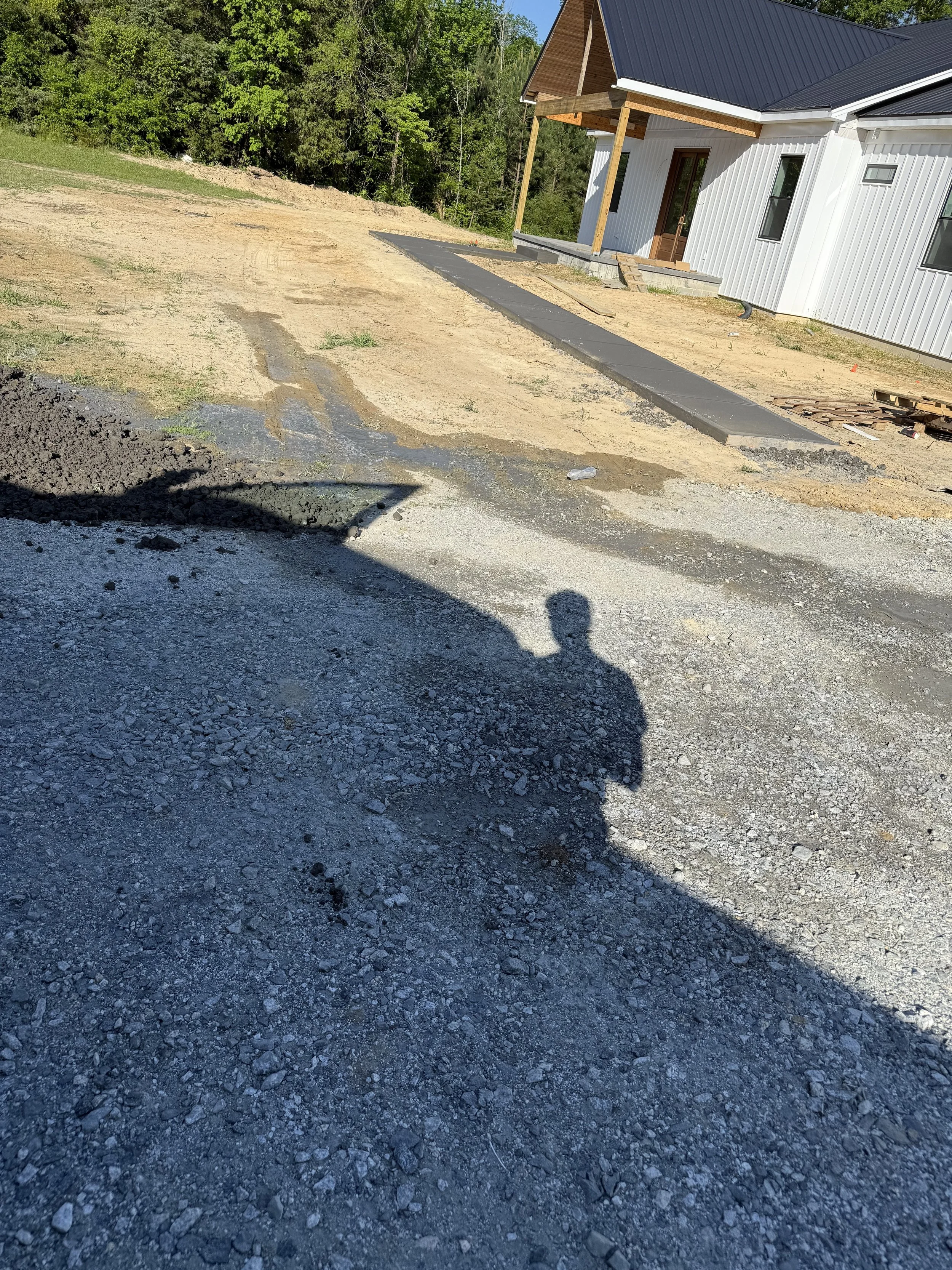 Construction site with newly poured concrete walkway in front of a modern house with white siding and a black metal roof, surrounded by dirt and gravel, with trees in the background.