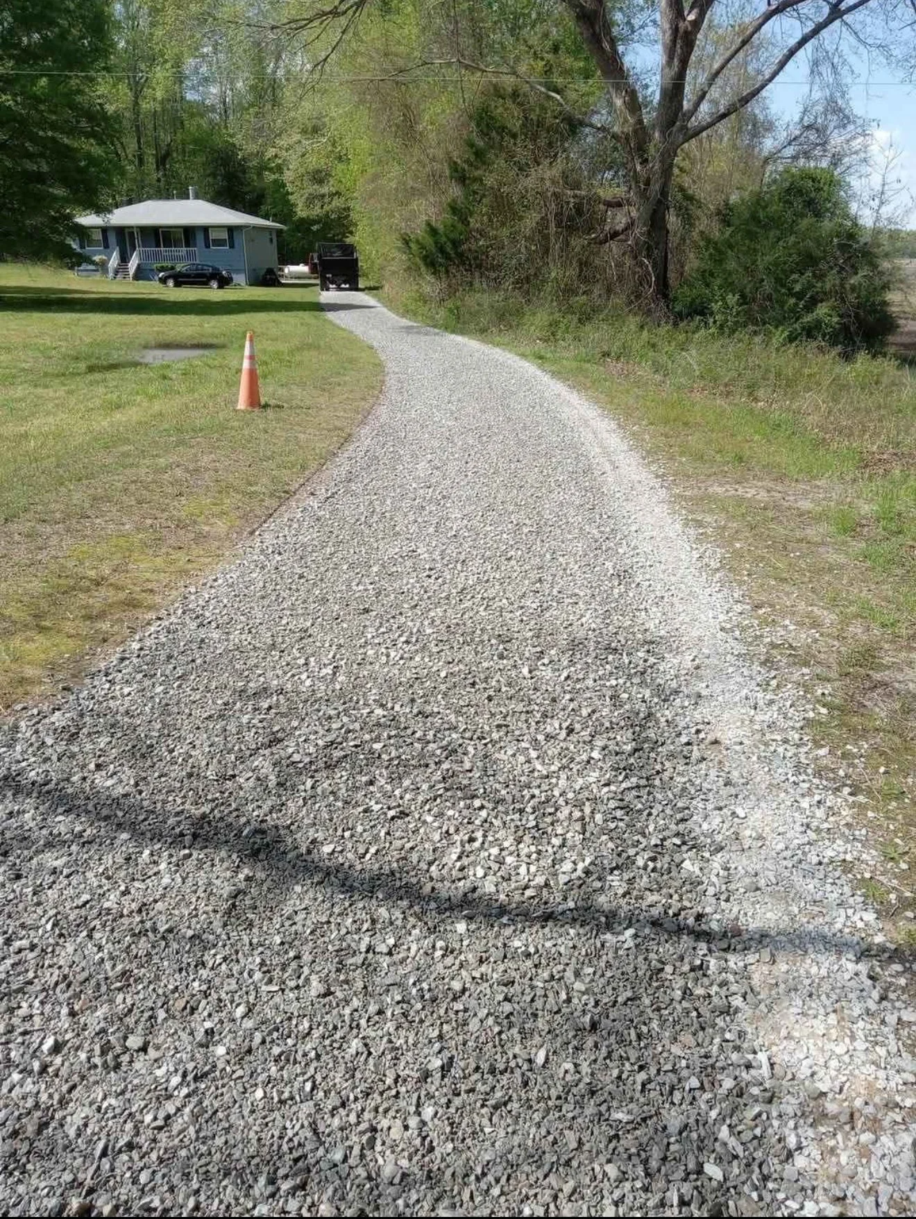 A gravel driveway leading to a house with a blue exterior, surrounded by green grass and trees. An orange traffic cone is on the grassy area near the driveway, and there's a parked black car and a truck near the house.