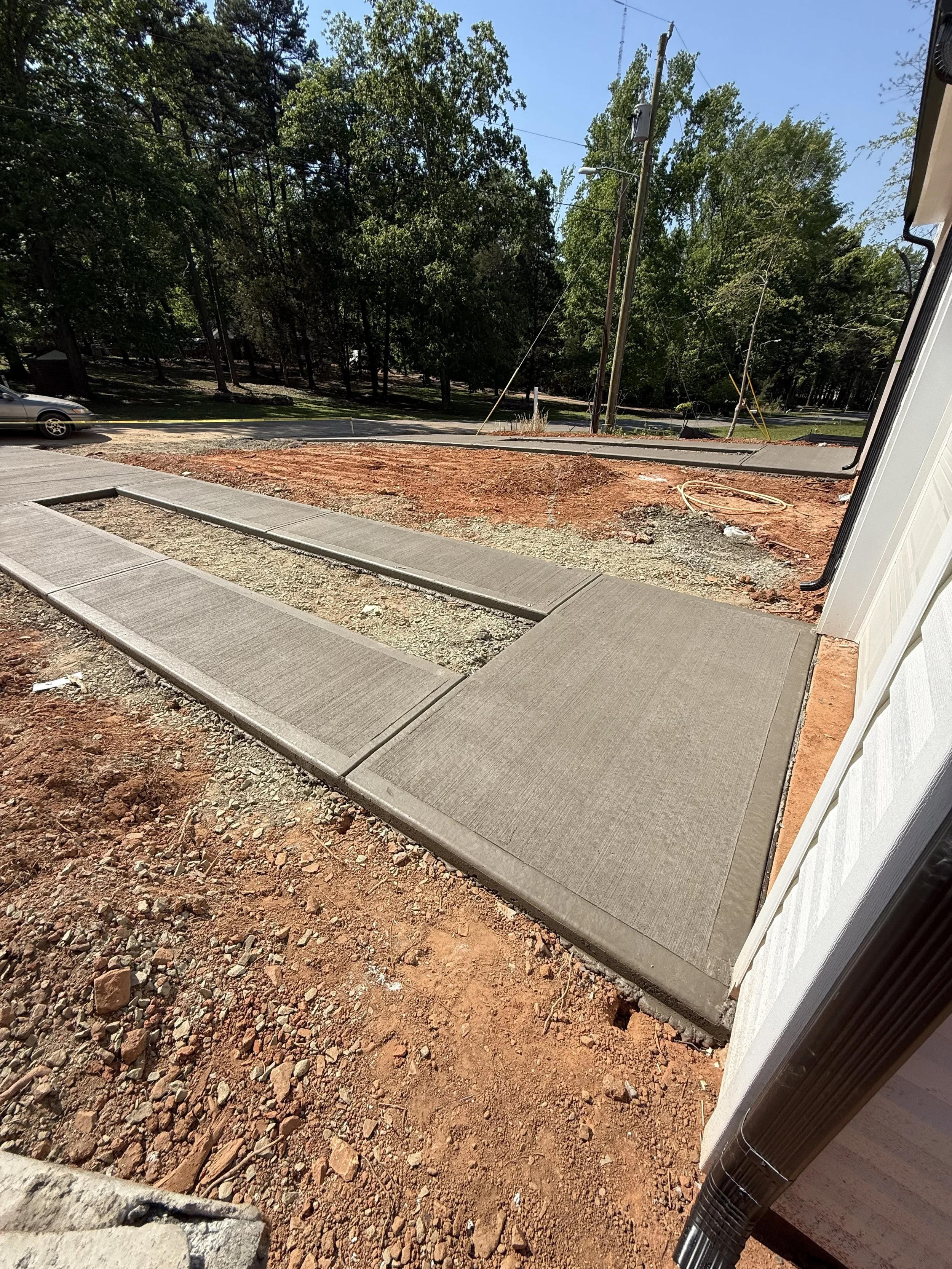 Newly poured cement sidewalks on a construction site with dirt and gravel surrounding them, and trees and power lines in the background.